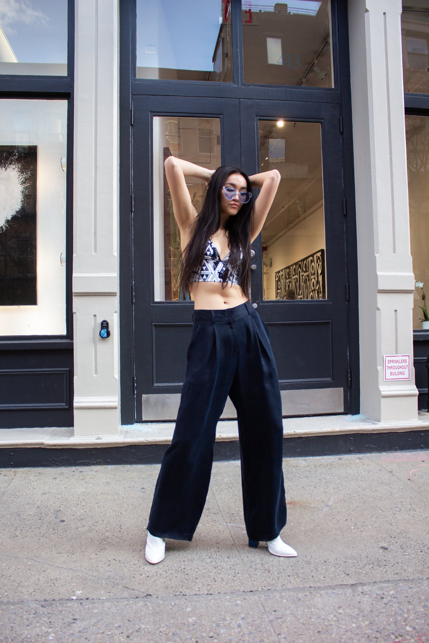A woman in sunglasses strolls down a city street, wearing a patterned crop top, Remi Navy Pants by Le Réussi®, and white shoes, with her hands tucked in her pockets.