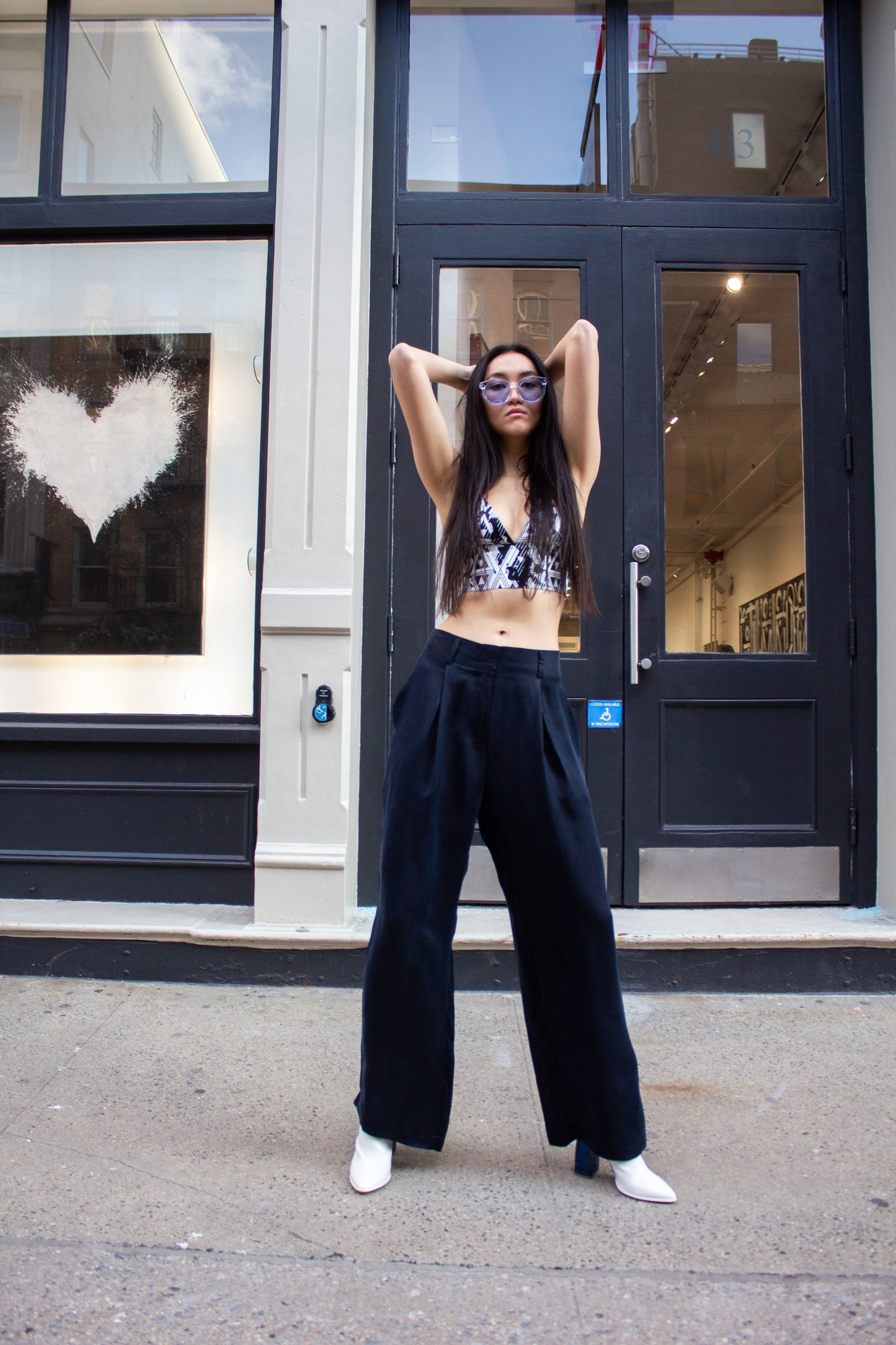 A woman in sunglasses strolls down a city street, wearing a patterned crop top, Remi Navy Pants by Le Réussi®, and white shoes, with her hands tucked in her pockets.