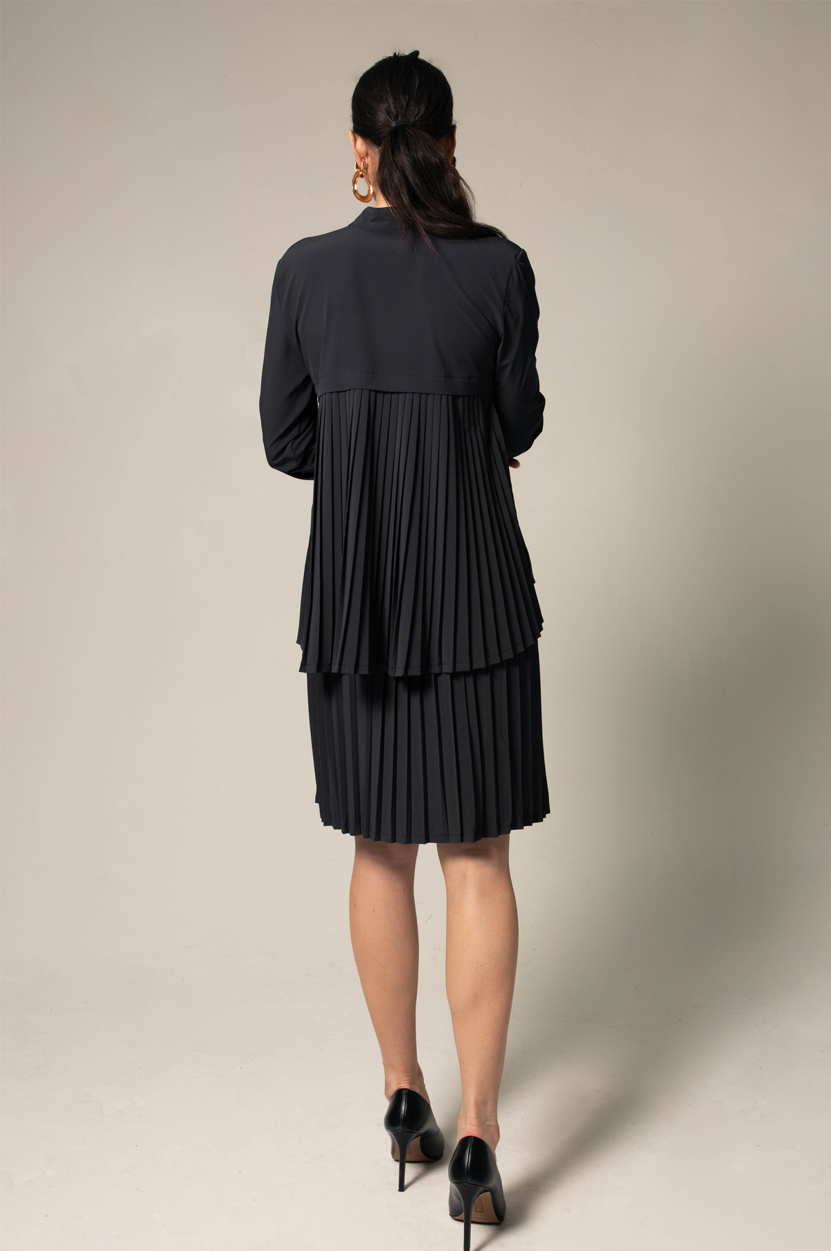 A woman wearing a black Stylish Pleated Blouse by Le Réussi® and high heels stands against a plain background, facing sideways.