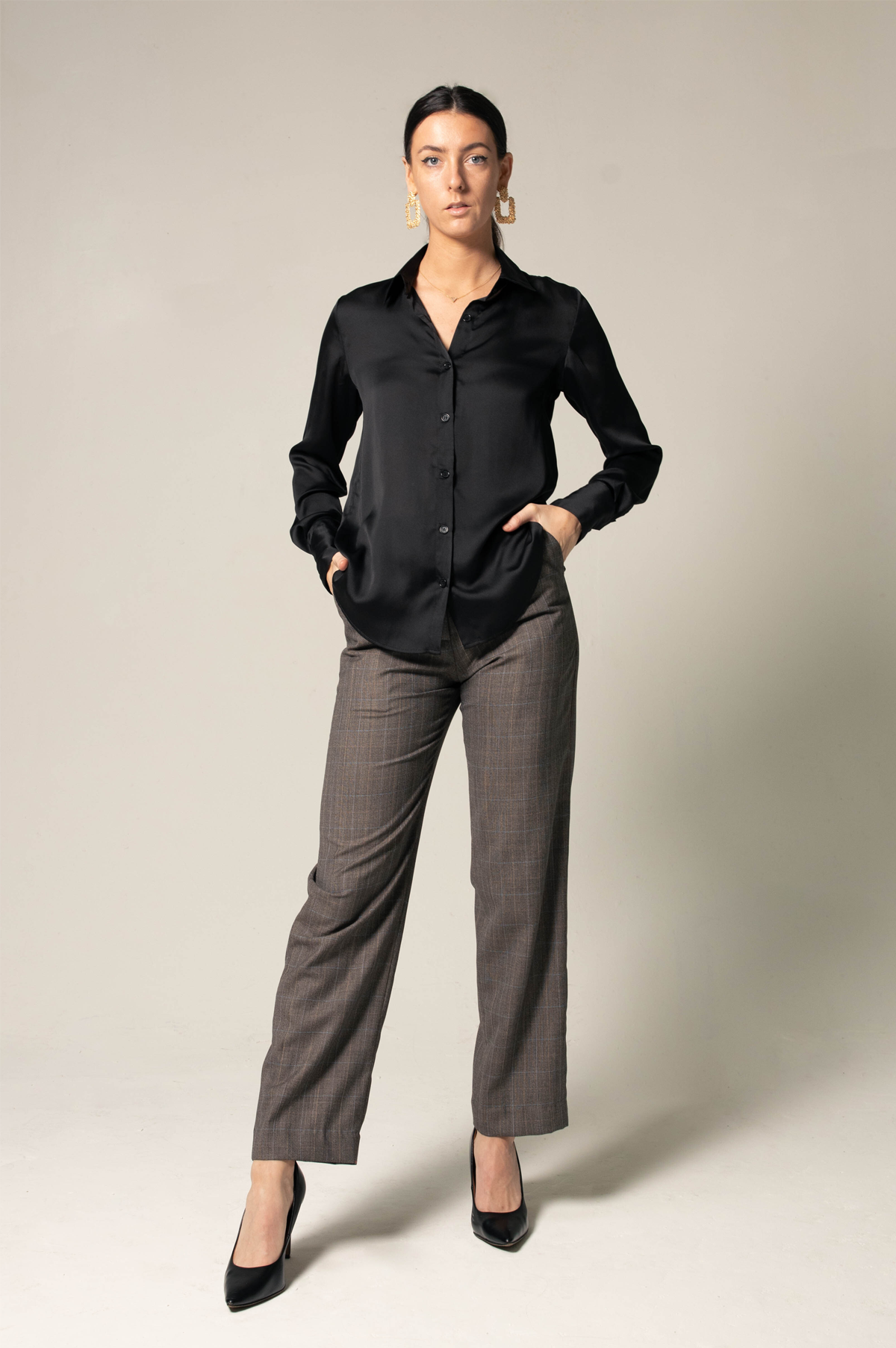 A person wearing the Elegance Silk Shirt in Black by Le Réussi®, paired with plaid trousers and black heels, stands with hands in pockets against a neutral background.