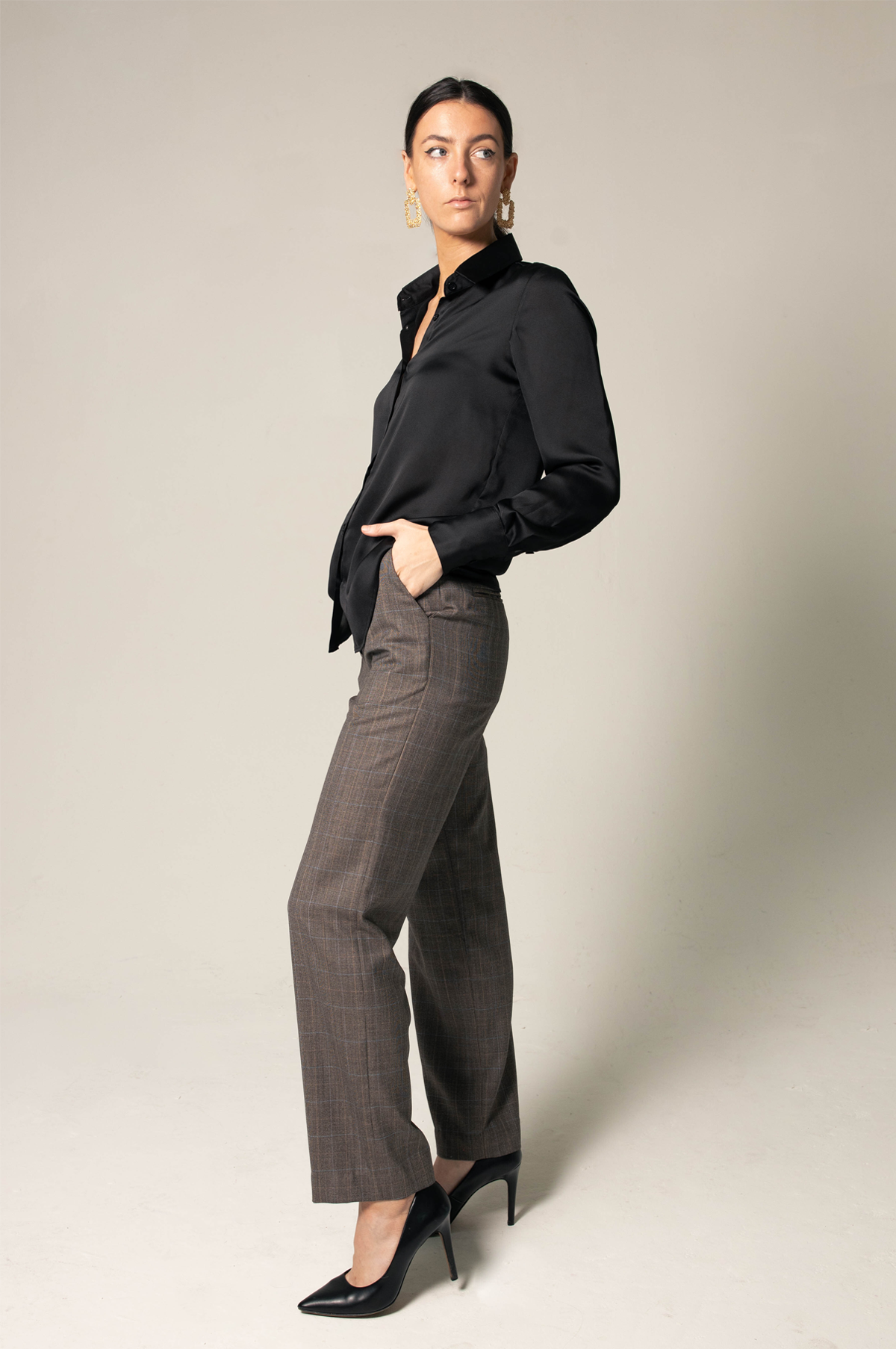 A person wearing the Elegance Silk Shirt in Black by Le Réussi®, paired with plaid trousers and black heels, stands with hands in pockets against a neutral background.