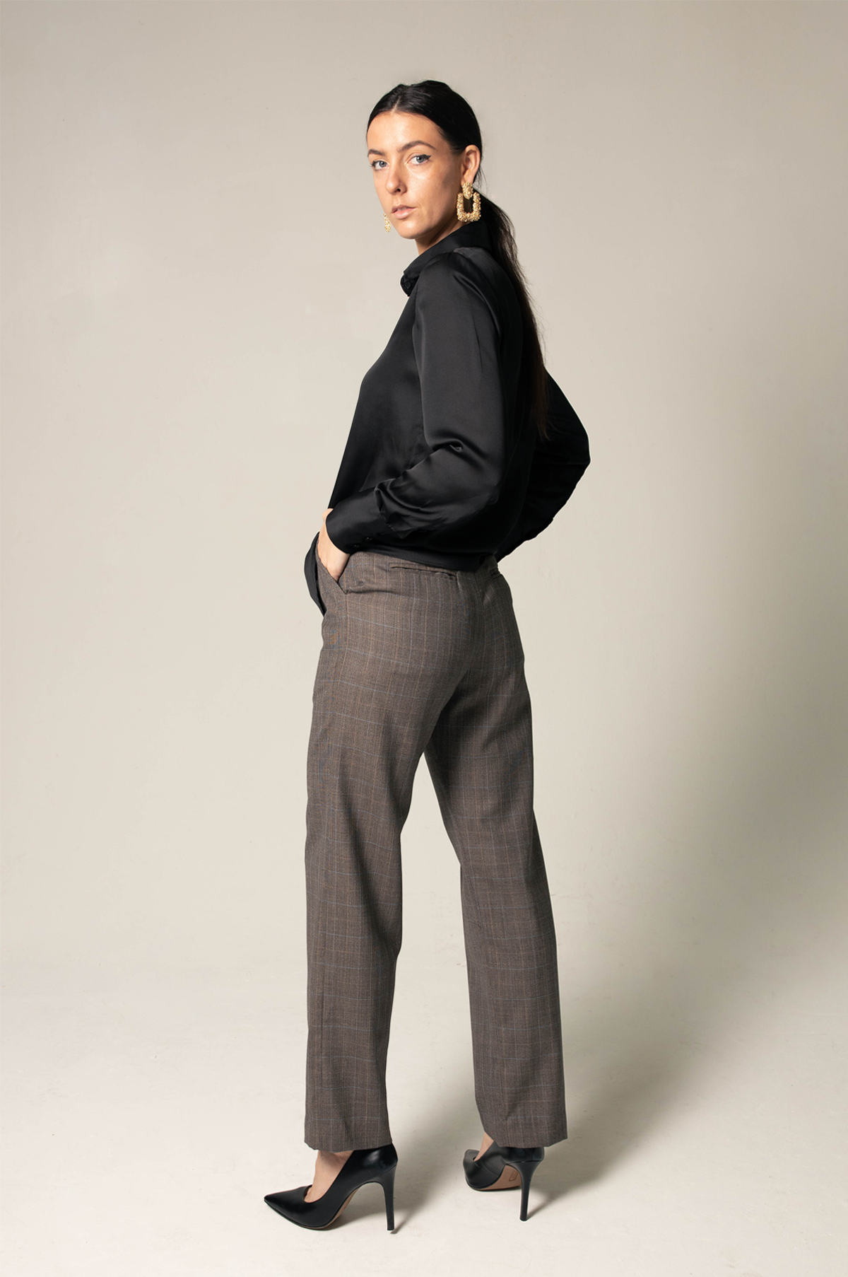 A person wearing the Elegance Silk Shirt in Black by Le Réussi®, paired with plaid trousers and black heels, stands with hands in pockets against a neutral background.