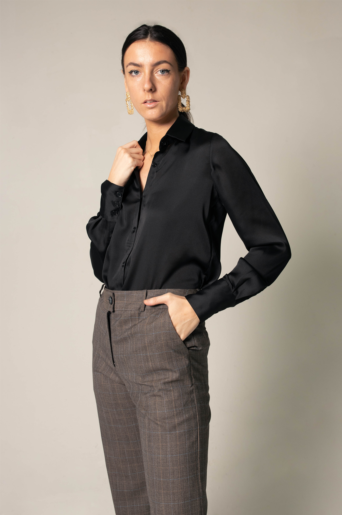 A person wearing the Elegance Silk Shirt in Black by Le Réussi®, paired with plaid trousers and black heels, stands with hands in pockets against a neutral background.