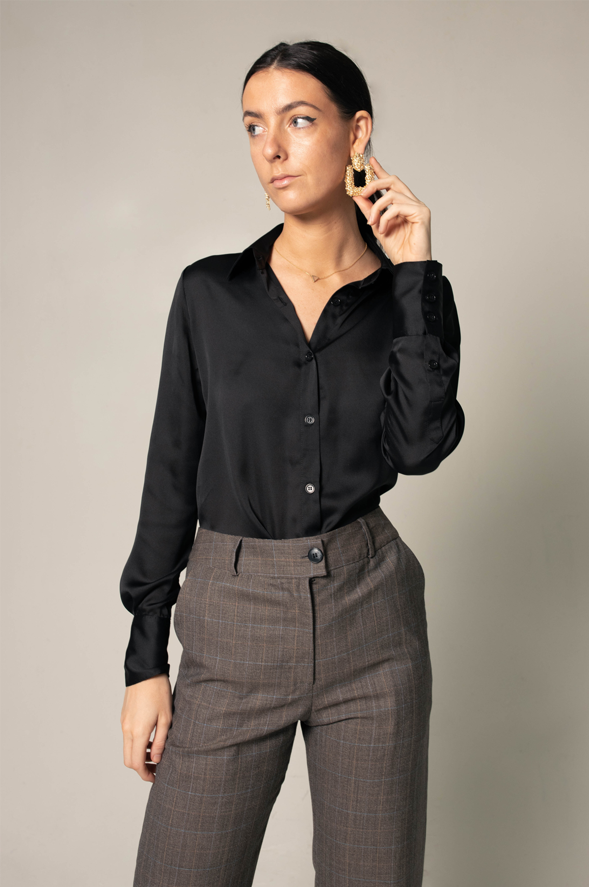 A person wearing the Elegance Silk Shirt in Black by Le Réussi®, paired with plaid trousers and black heels, stands with hands in pockets against a neutral background.