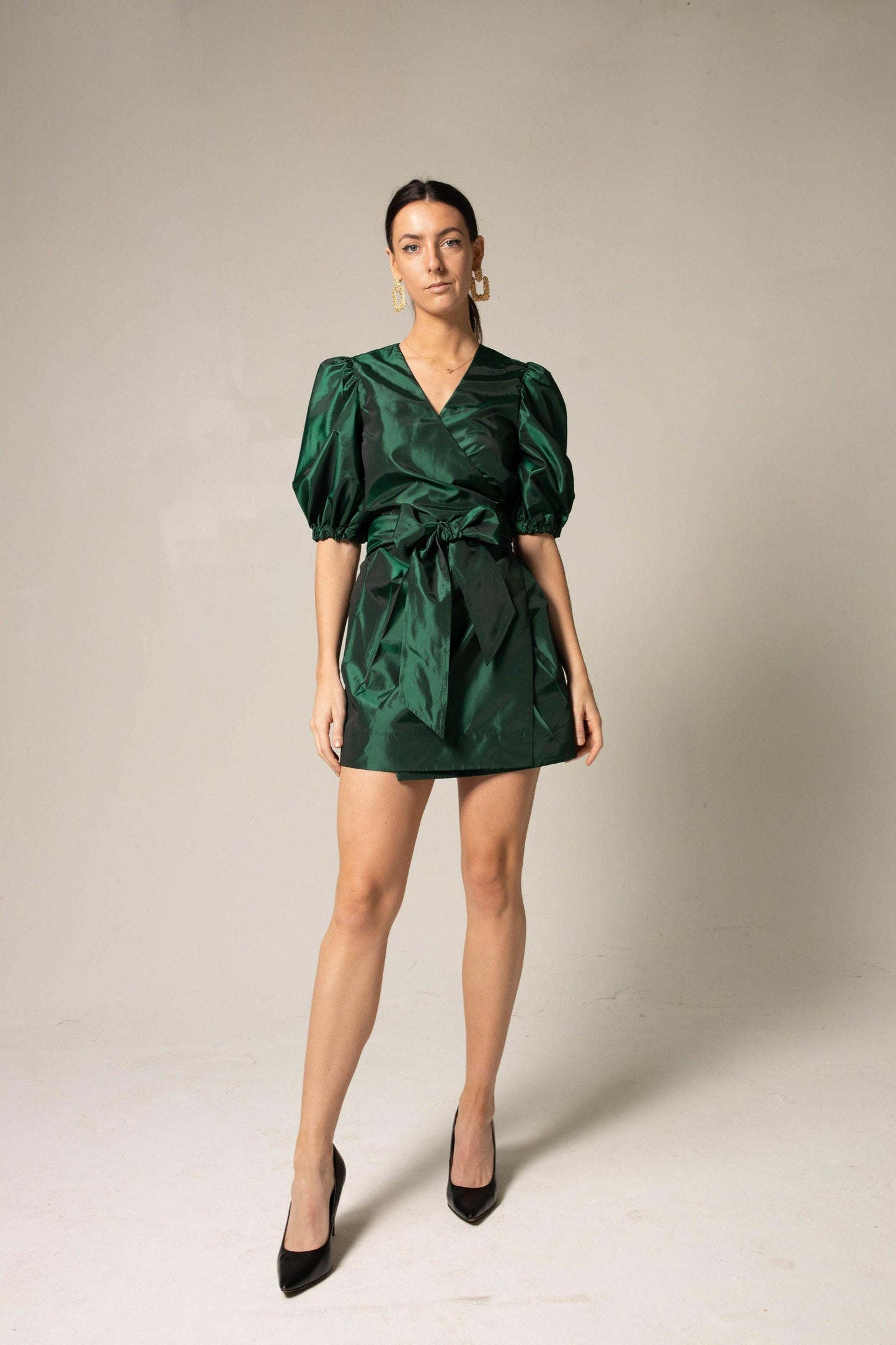 A woman in the Glimmer Green Wrap Dress by Le Réussi® stands against a neutral background, wearing black heels and gold earrings.