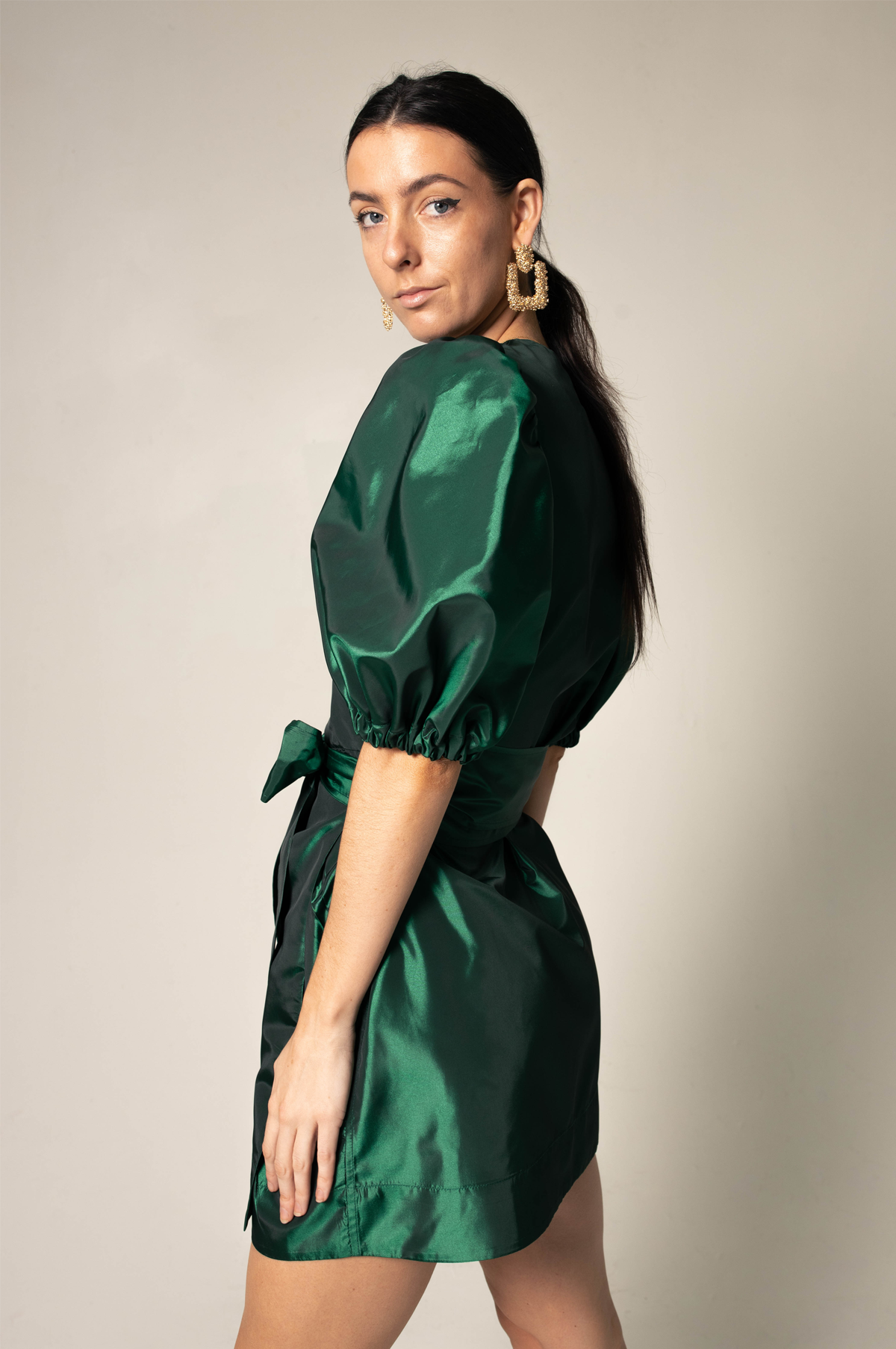 A woman in the Glimmer Green Wrap Dress by Le Réussi® stands against a neutral background, wearing black heels and gold earrings.