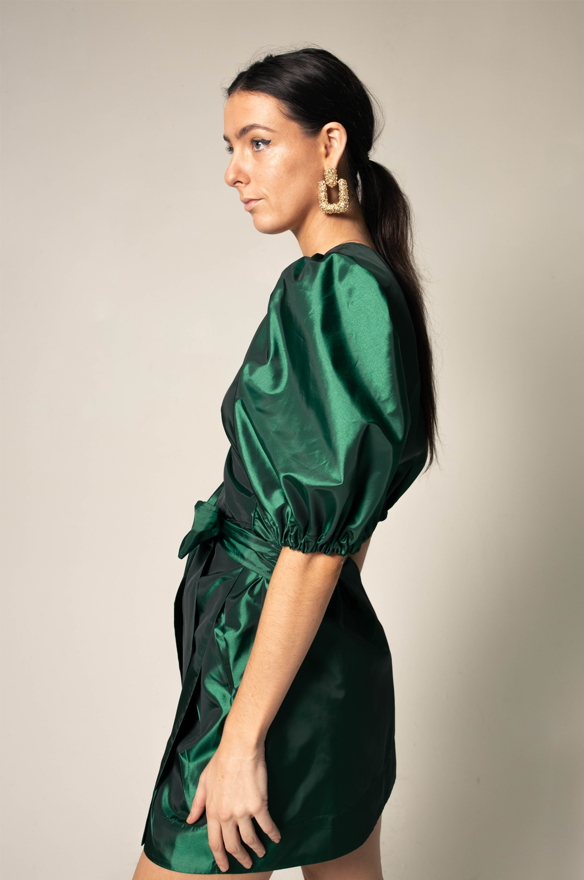 A woman in the Glimmer Green Wrap Dress by Le Réussi® stands against a neutral background, wearing black heels and gold earrings.