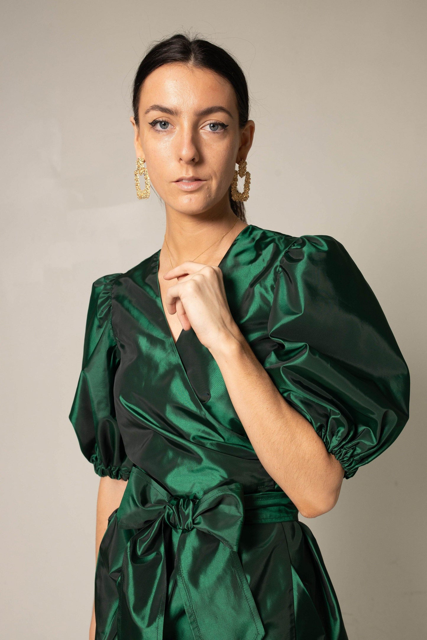 A woman in the Glimmer Green Wrap Dress by Le Réussi® stands against a neutral background, wearing black heels and gold earrings.