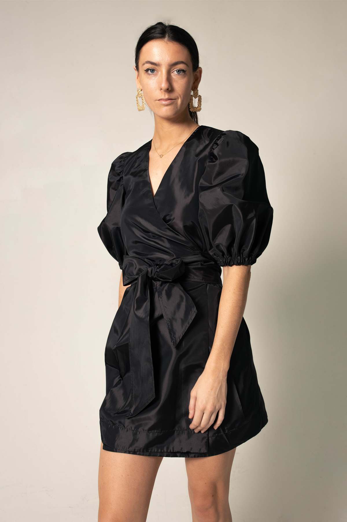 A person stands against a plain background wearing Le Réussi®s Black Lusso Wrap Dress paired with large gold hoop earrings.