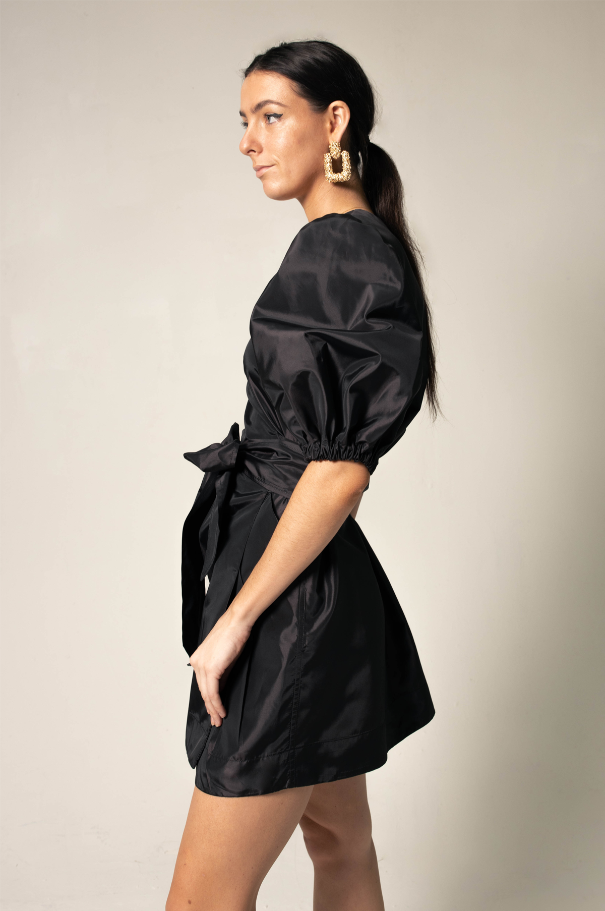 A person stands against a plain background wearing Le Réussi®s Black Lusso Wrap Dress paired with large gold hoop earrings.