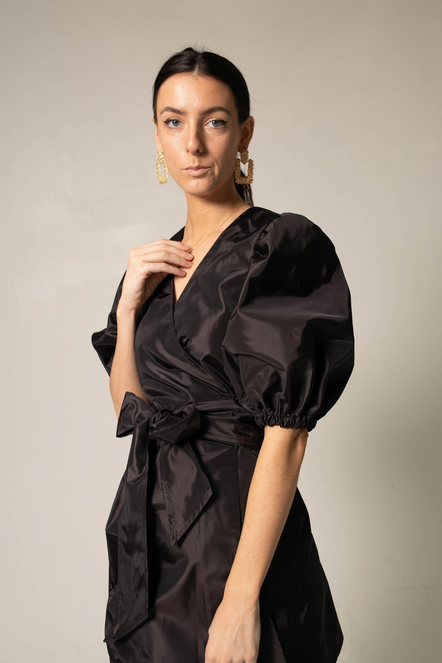 A person stands against a plain background wearing Le Réussi®s Black Lusso Wrap Dress paired with large gold hoop earrings.