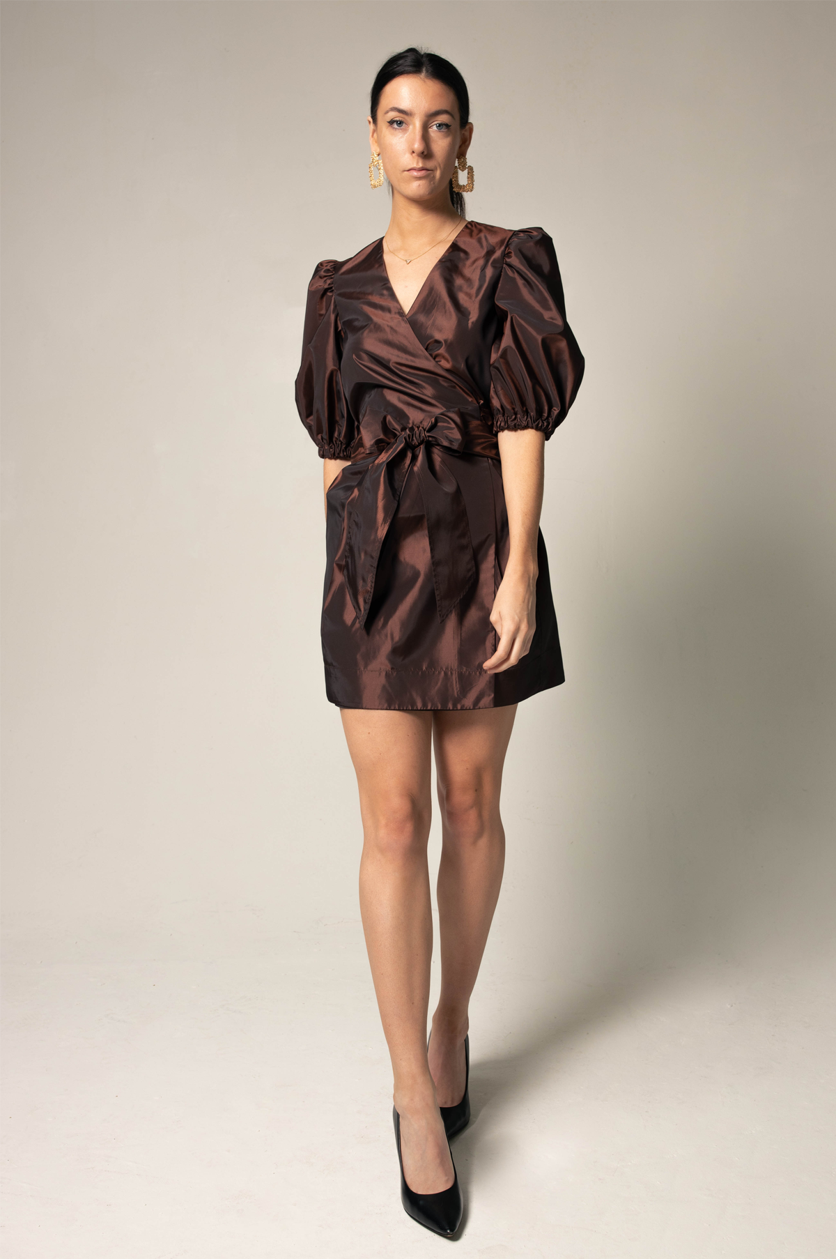 A person in a Power Bordeaux Red Wrap Dress by Le Réussi® stands against a plain background, featuring satin fabric with puff sleeves and a bow detail.