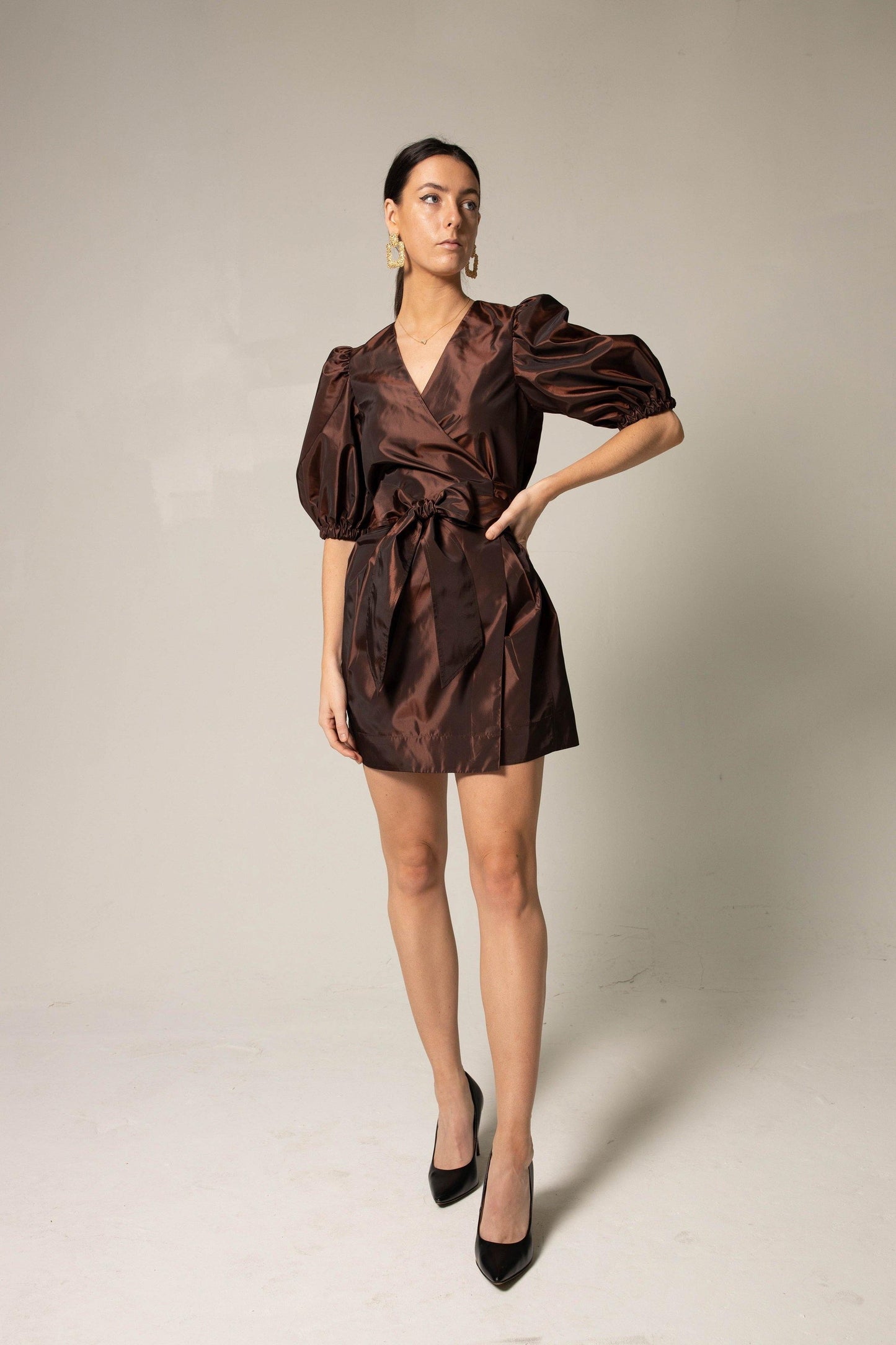 A person in a Power Bordeaux Red Wrap Dress by Le Réussi® stands against a plain background, featuring satin fabric with puff sleeves and a bow detail.