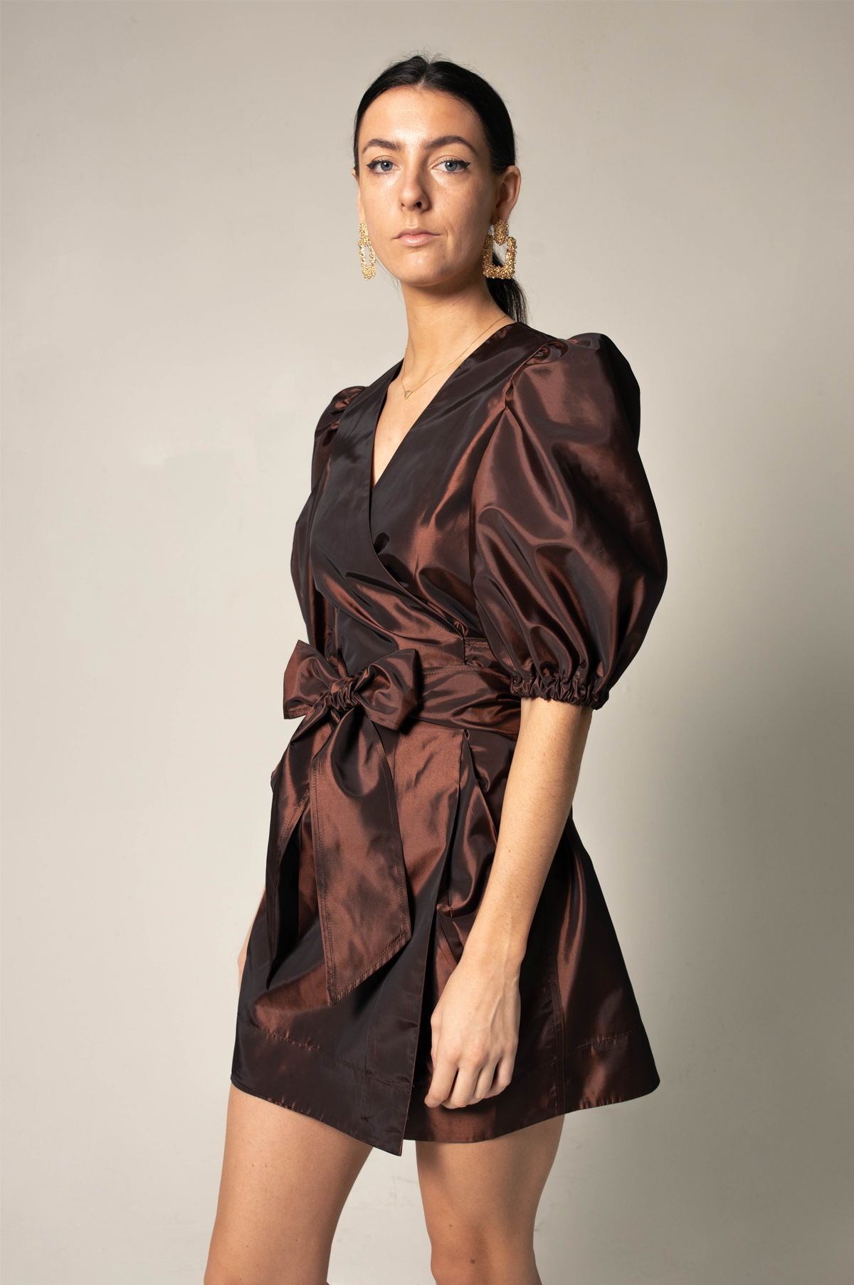 A person in a Power Bordeaux Red Wrap Dress by Le Réussi® stands against a plain background, featuring satin fabric with puff sleeves and a bow detail.