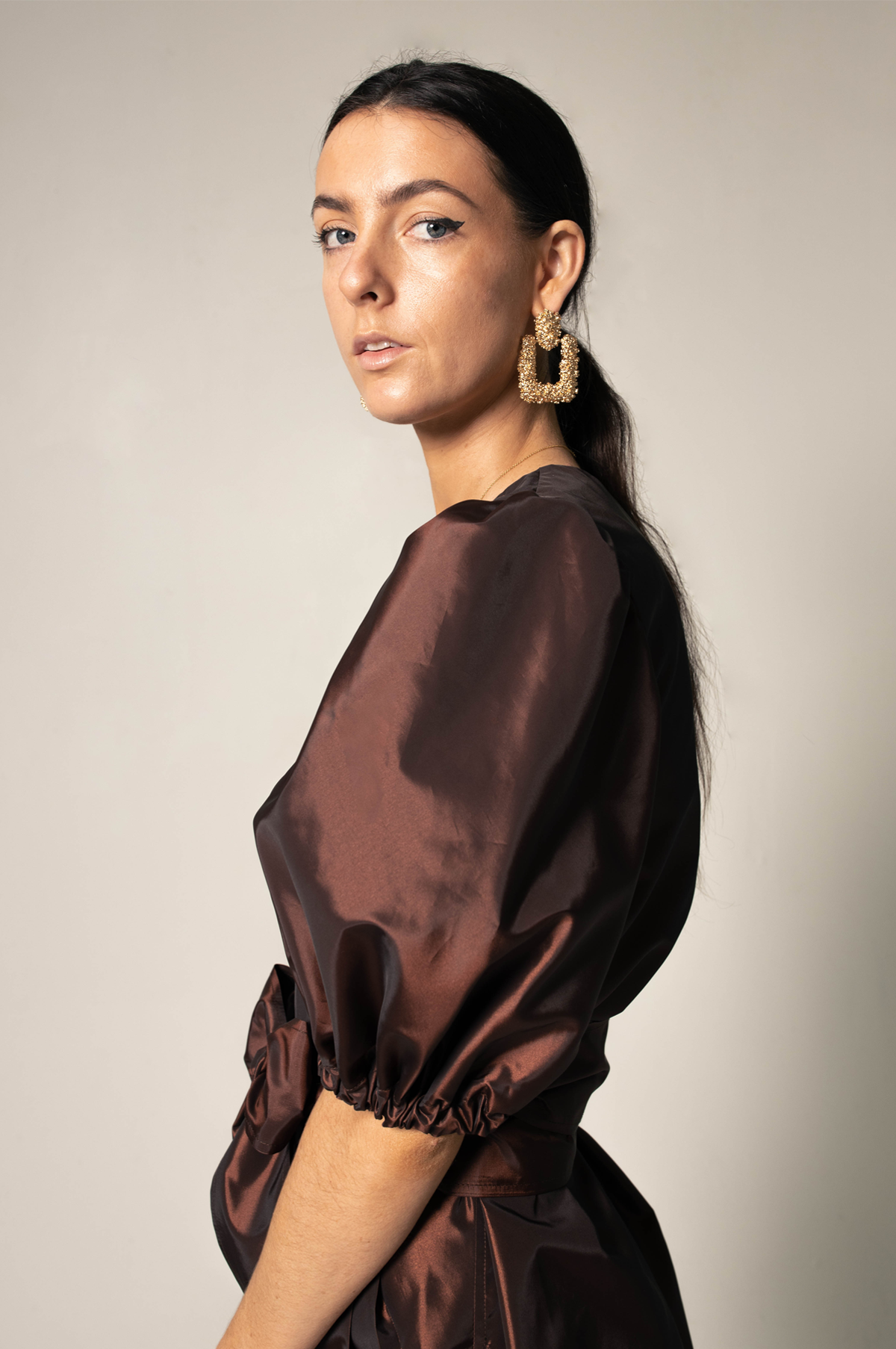 A person in a Power Bordeaux Red Wrap Dress by Le Réussi® stands against a plain background, featuring satin fabric with puff sleeves and a bow detail.
