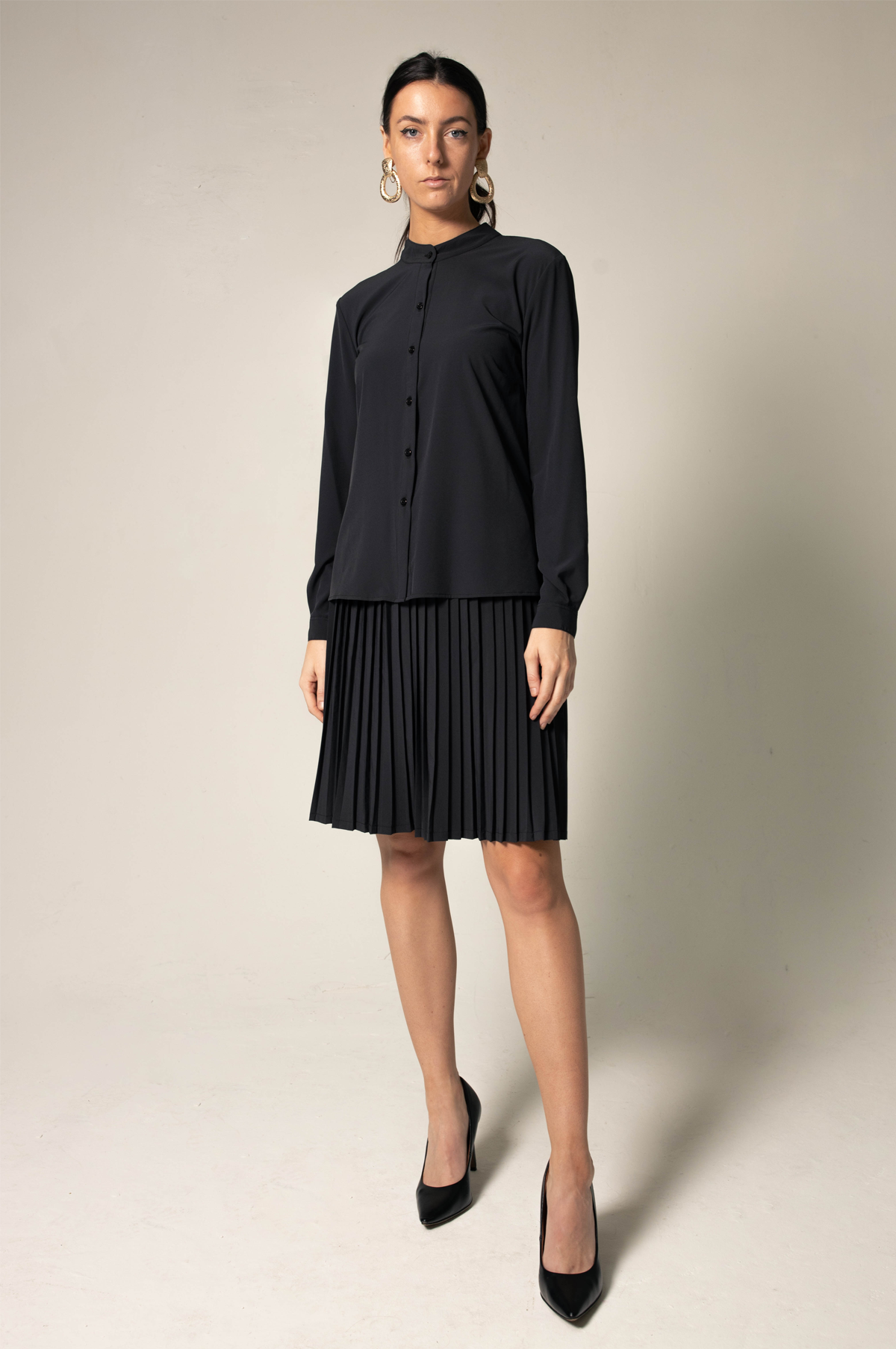 A woman wearing a black Stylish Pleated Blouse by Le Réussi® and high heels stands against a plain background, facing sideways.