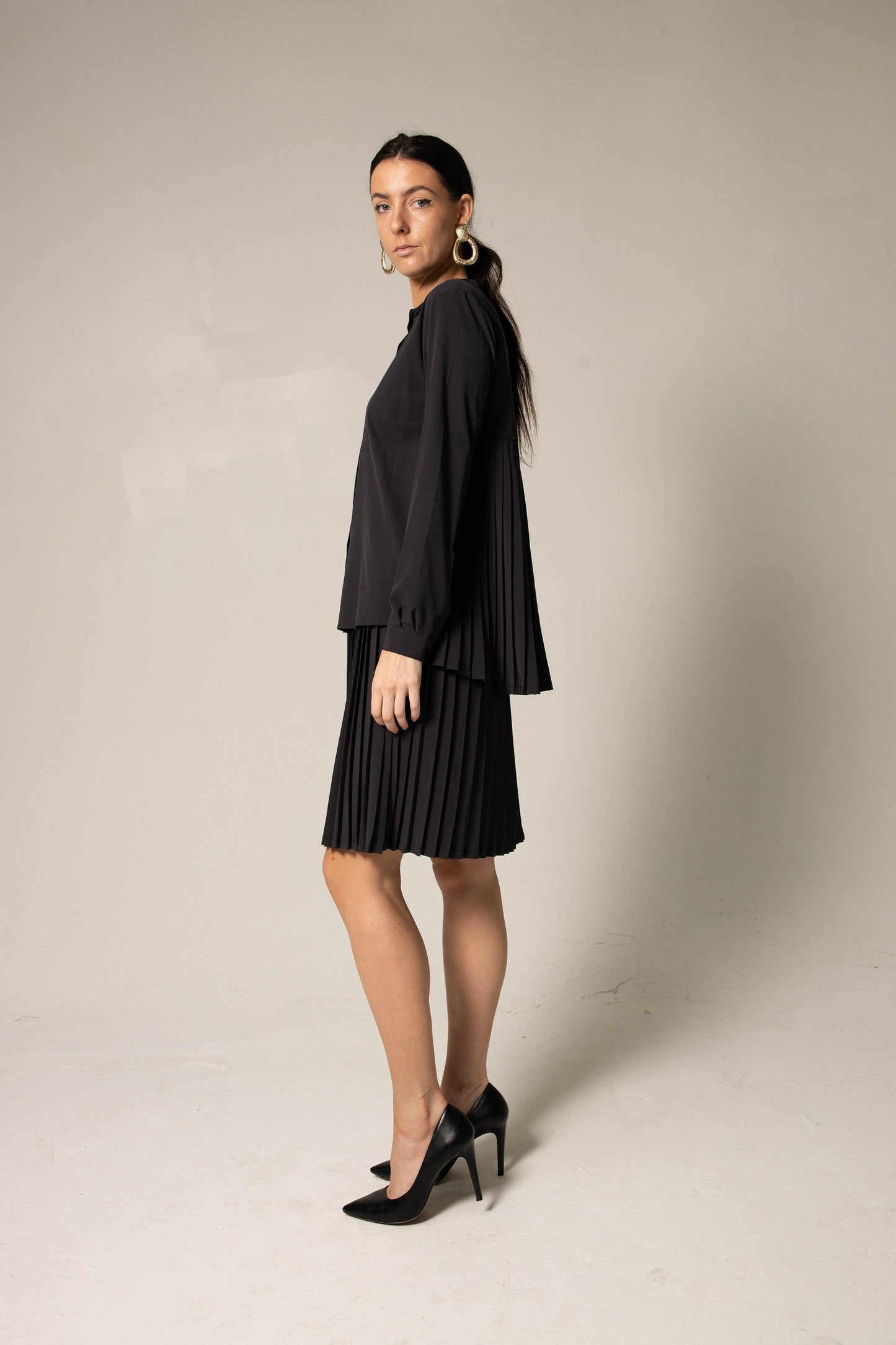 A woman in a Stylish Pleated Blouse in Black by Le Réussi® and heels poses against a plain background, accessorized with large hoop earrings.