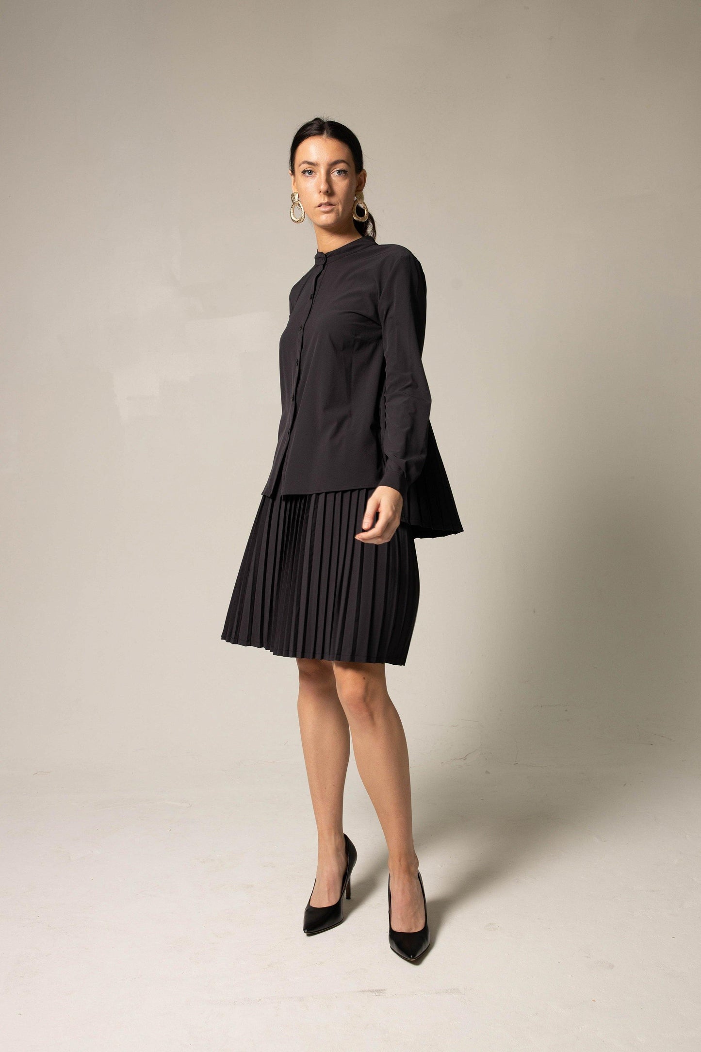A woman in a Stylish Pleated Blouse in Black by Le Réussi® and heels poses against a plain background, accessorized with large hoop earrings.