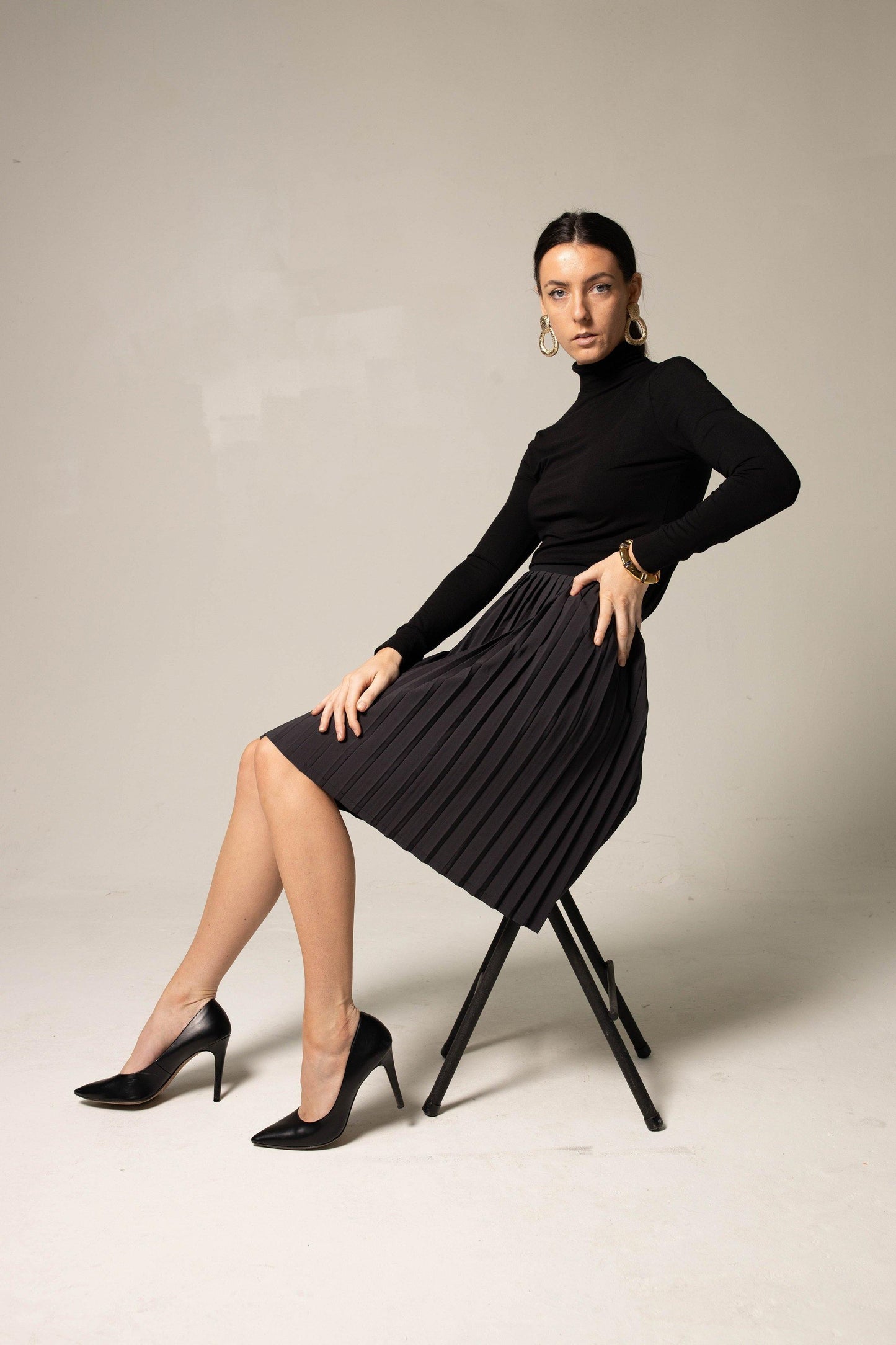 Sporting a black turtleneck, Le Réussi® Classy Pleated Skirt, and heels, a person stands in a side pose against a plain background.