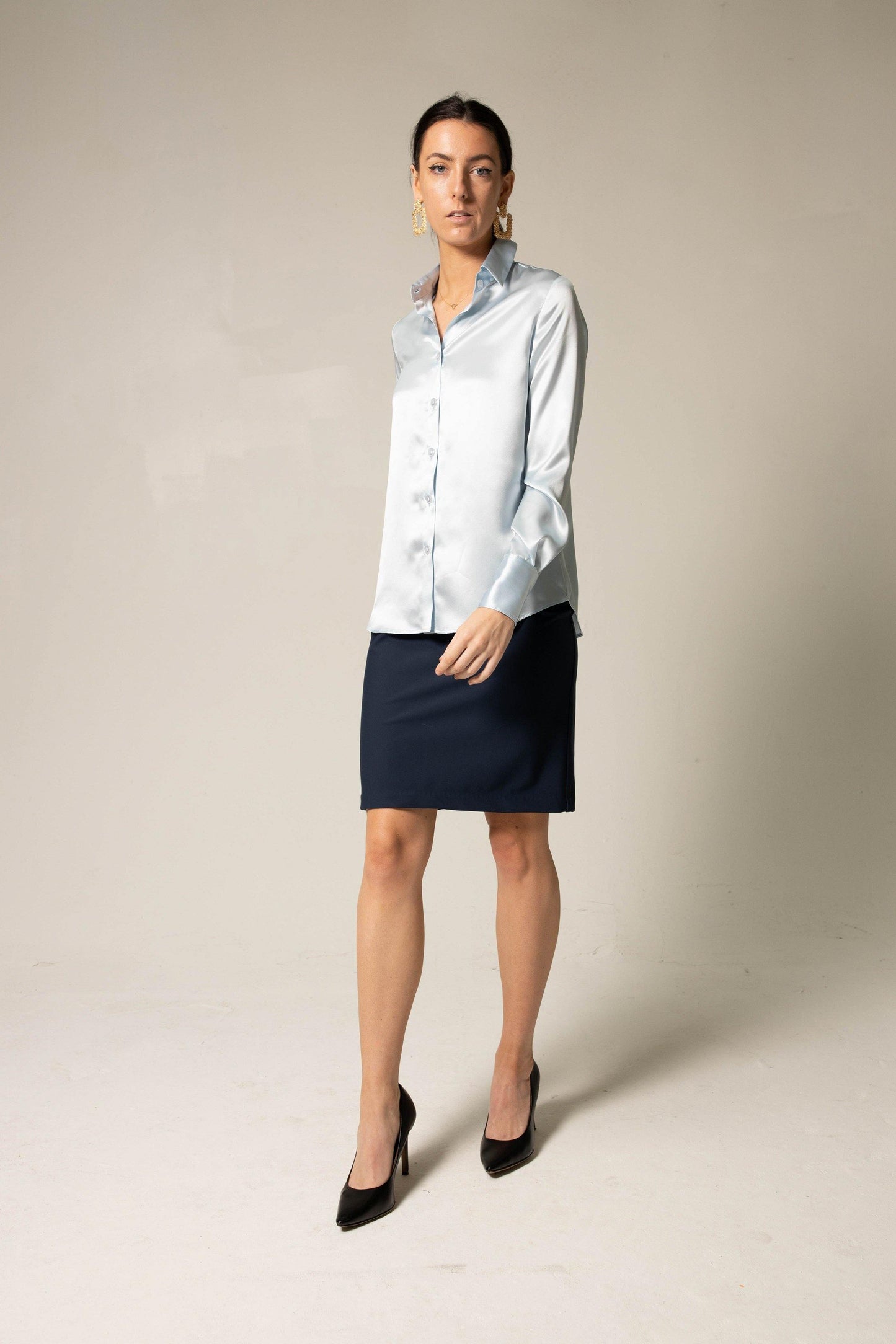 A woman in a light blue satin blouse and Navy Womens Straight Skirt by Le Réussi® stands against a plain background, wearing black heels and gold earrings.