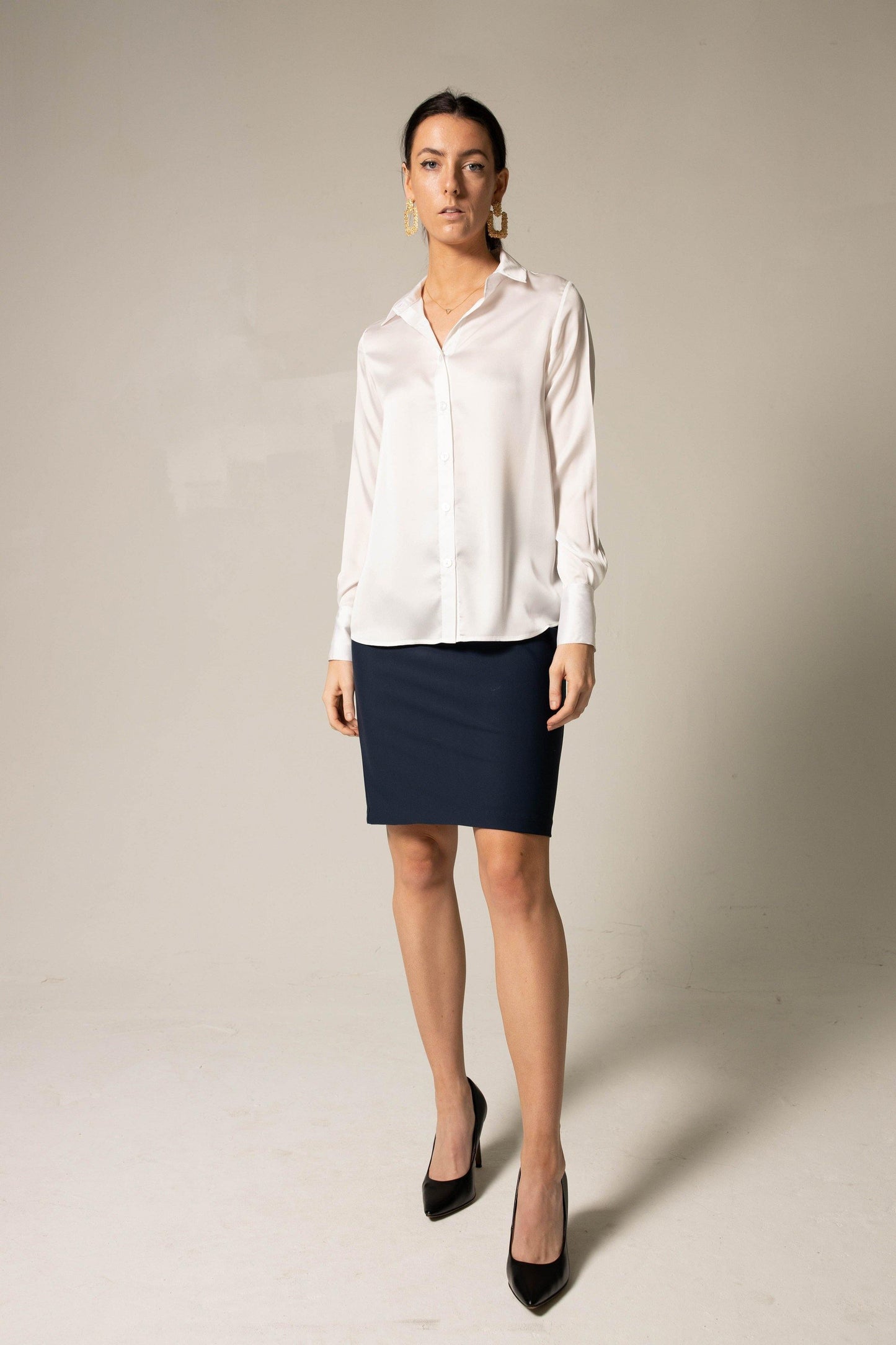A woman poses against a neutral background wearing the Elegance Silk Shirt in White by Le Réussi®, paired with a dark skirt.