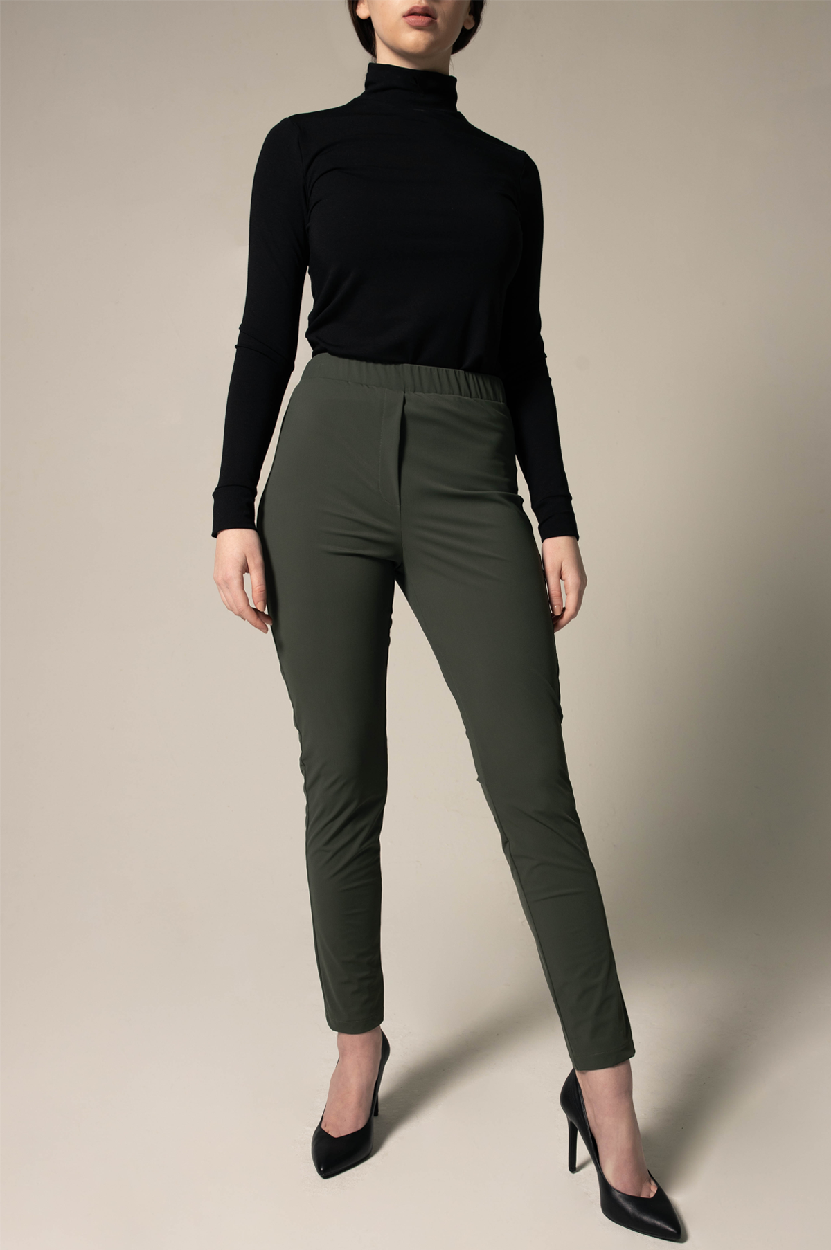 A person wearing a black turtleneck, Le Réussi® Olive Skinny Pants Womens Trousers, and black heels stands against a plain background.