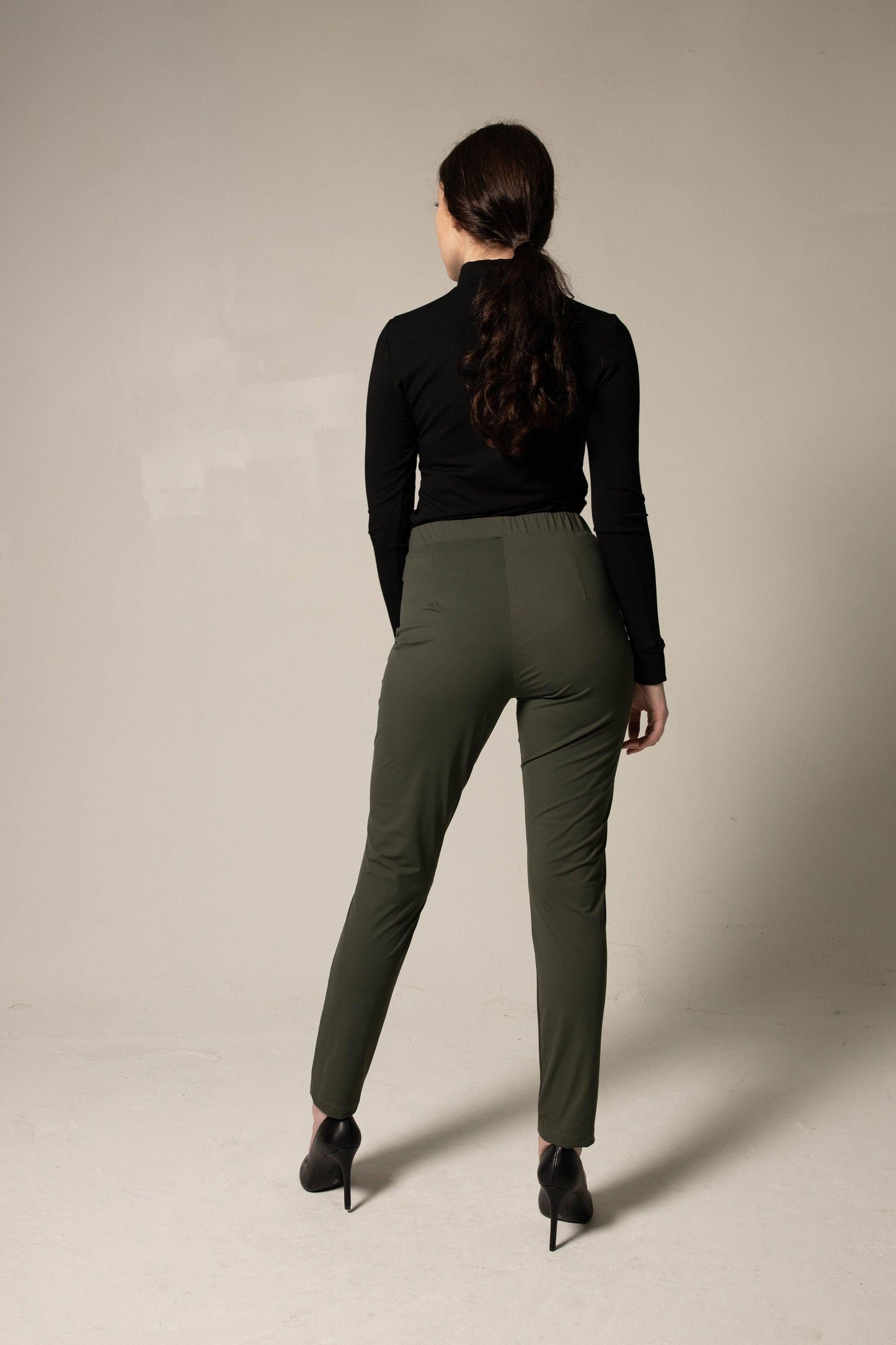A person wearing a black turtleneck, Le Réussi® Olive Skinny Pants Womens Trousers, and black heels stands against a plain background.