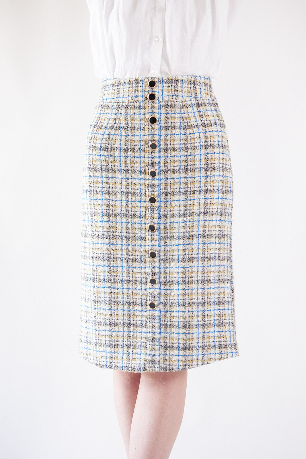 A woman stands against a white background in a Le Réussi® Power Woman- Eye Catching Tweed Skirt, paired with a white blouse, ankle boots, and a pearl necklace.