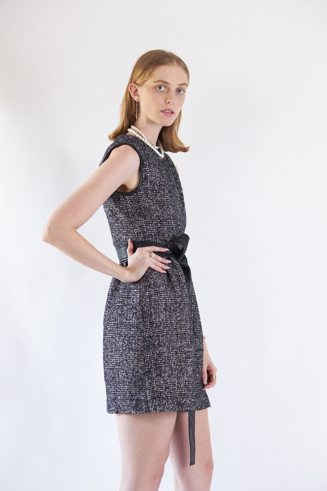 A person standing against a white background wears the Boss Woman Tweed Dress by Le Réussi®, accessorized with layered necklaces, a black belt, and black ankle boots.