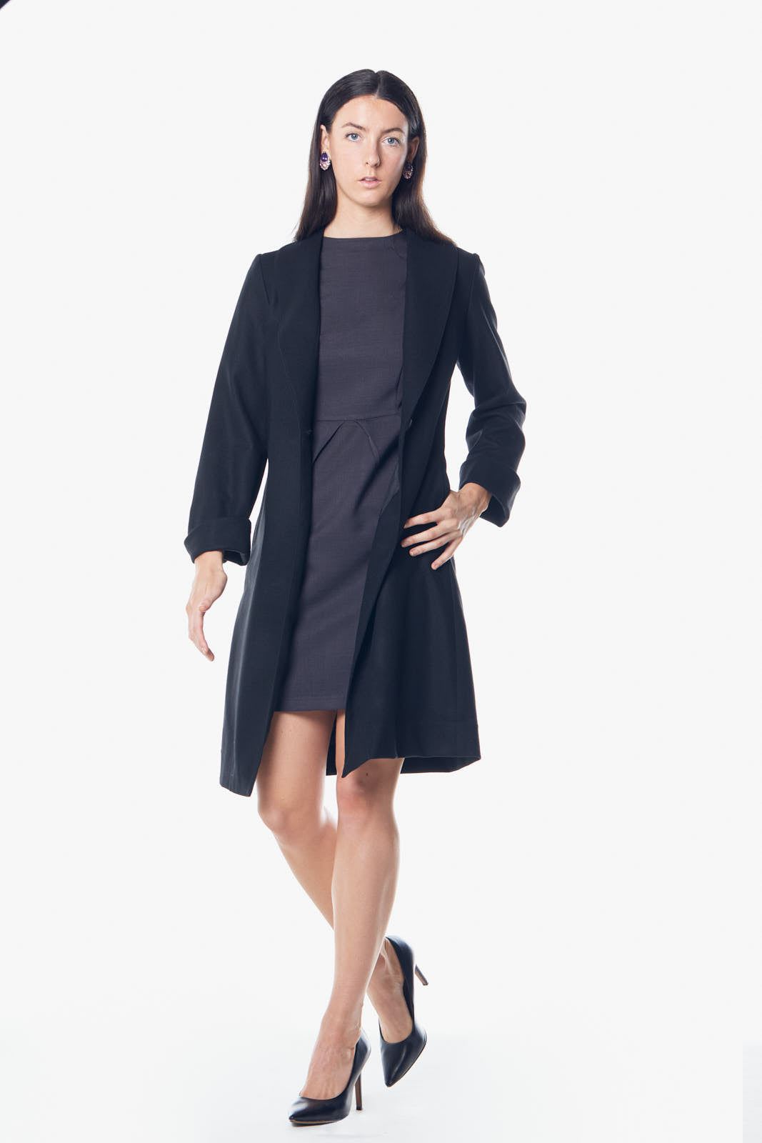 A woman stands against a white background wearing Le Réussi’s black Power Woman Worsted Flannel Long Jacket, paired with a dress and high heels.