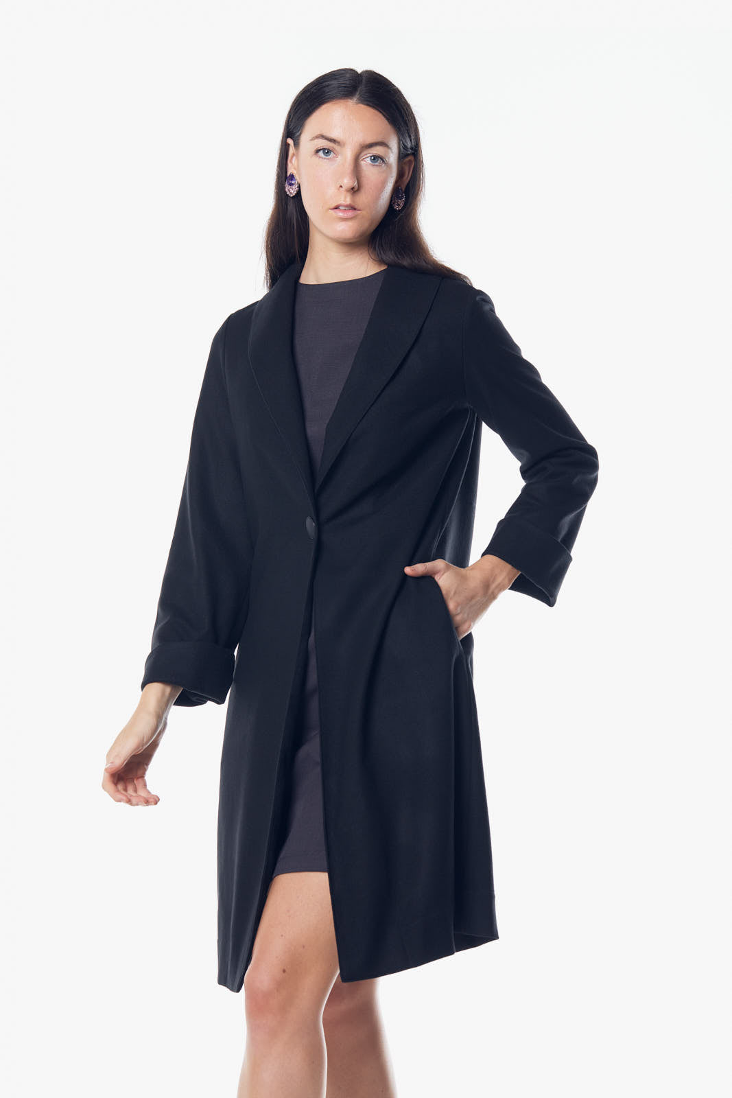 A woman stands against a white background wearing Le Réussi’s black Power Woman Worsted Flannel Long Jacket, paired with a dress and high heels.