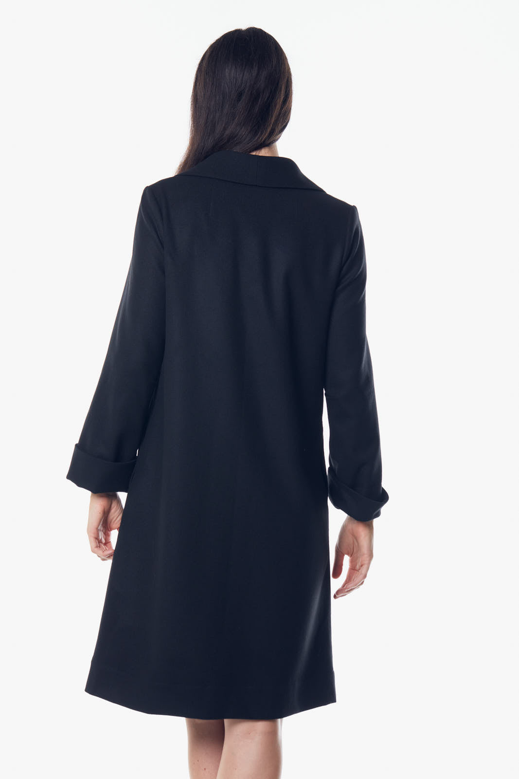 A woman stands against a white background wearing Le Réussi’s black Power Woman Worsted Flannel Long Jacket, paired with a dress and high heels.