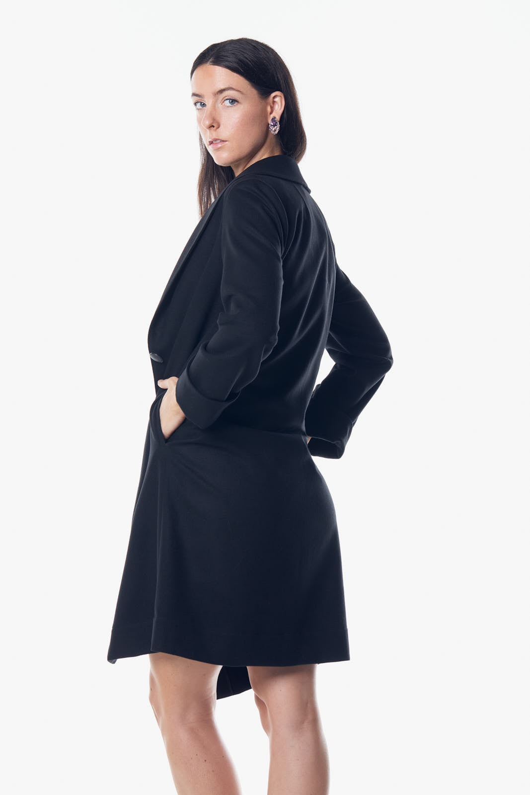 A woman stands against a white background wearing Le Réussi’s black Power Woman Worsted Flannel Long Jacket, paired with a dress and high heels.