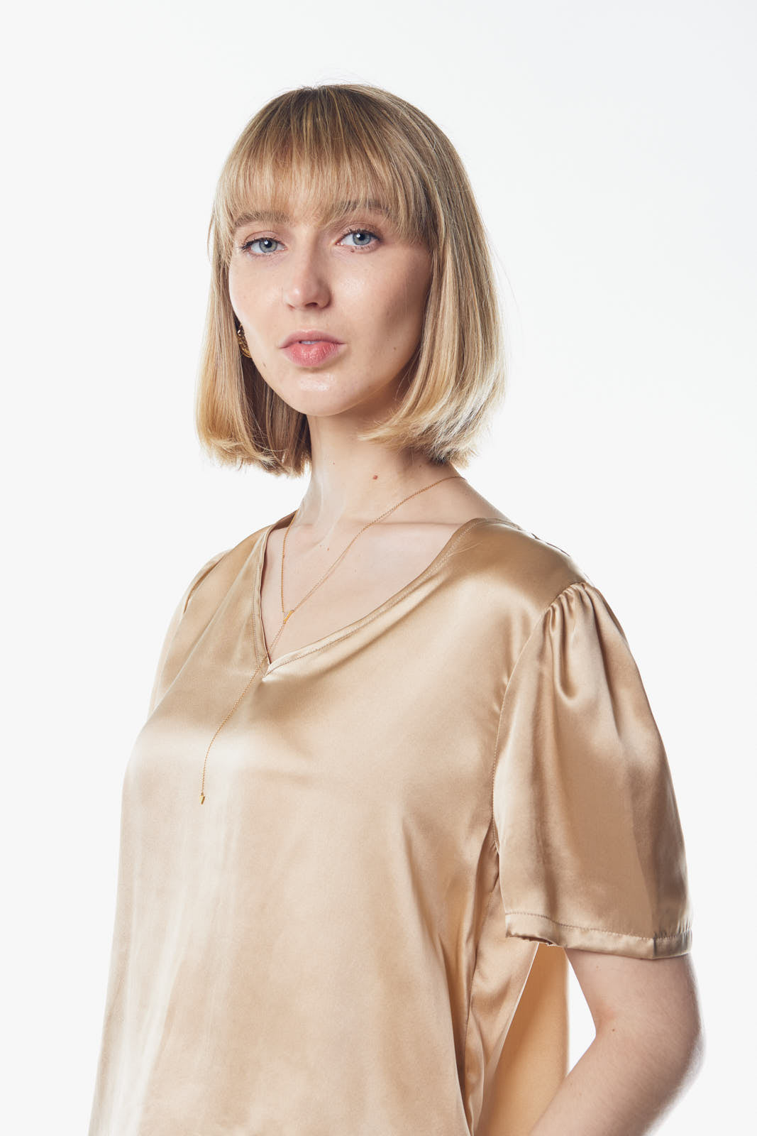 A person wearing the Le Réussi® Champagne V-Neck Blouse and dark pants stands against a white background, hands in pockets.