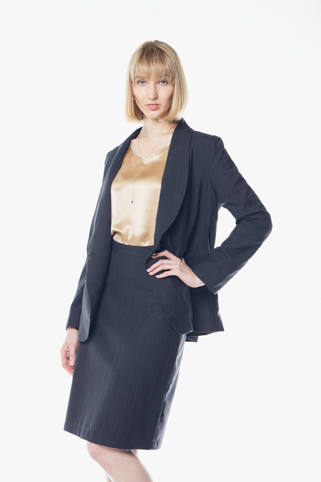 A person with short blond hair poses confidently in a beige top, skirt, and the Tailor Wool Dark Grey Blazer by Le Réussi® against a white background.