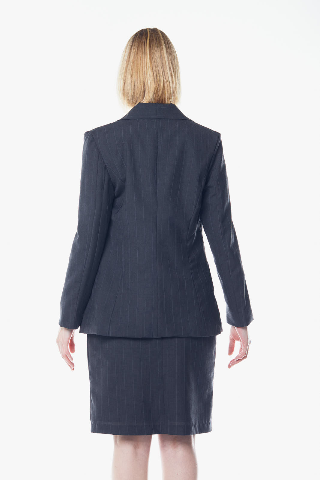 A person with short blond hair poses confidently in a beige top, skirt, and the Tailor Wool Dark Grey Blazer by Le Réussi® against a white background.