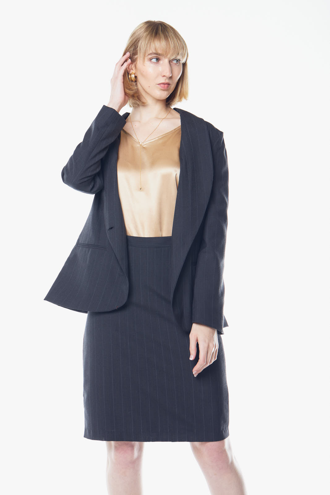A person with short blond hair poses confidently in a beige top, skirt, and the Tailor Wool Dark Grey Blazer by Le Réussi® against a white background.