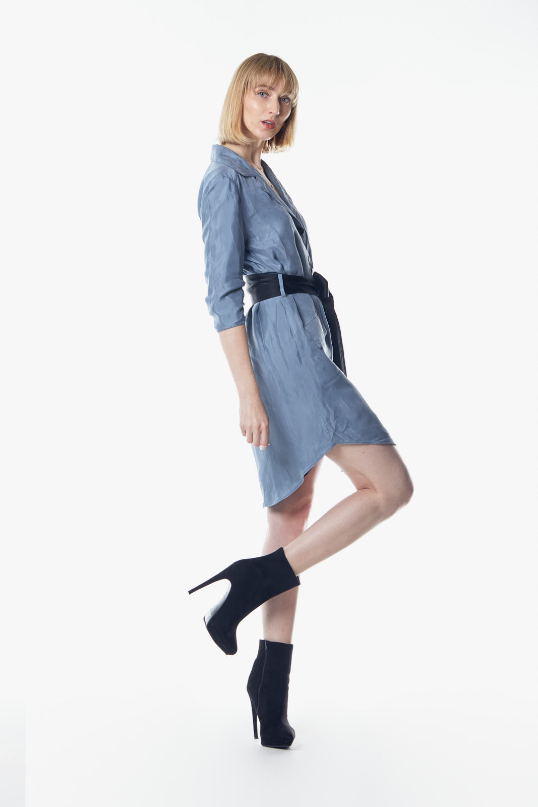 A person stands against a plain background, wearing the Cielo Blue Blazer Dress by Le Réussi®, accented with a black belt and paired with black ankle boots.