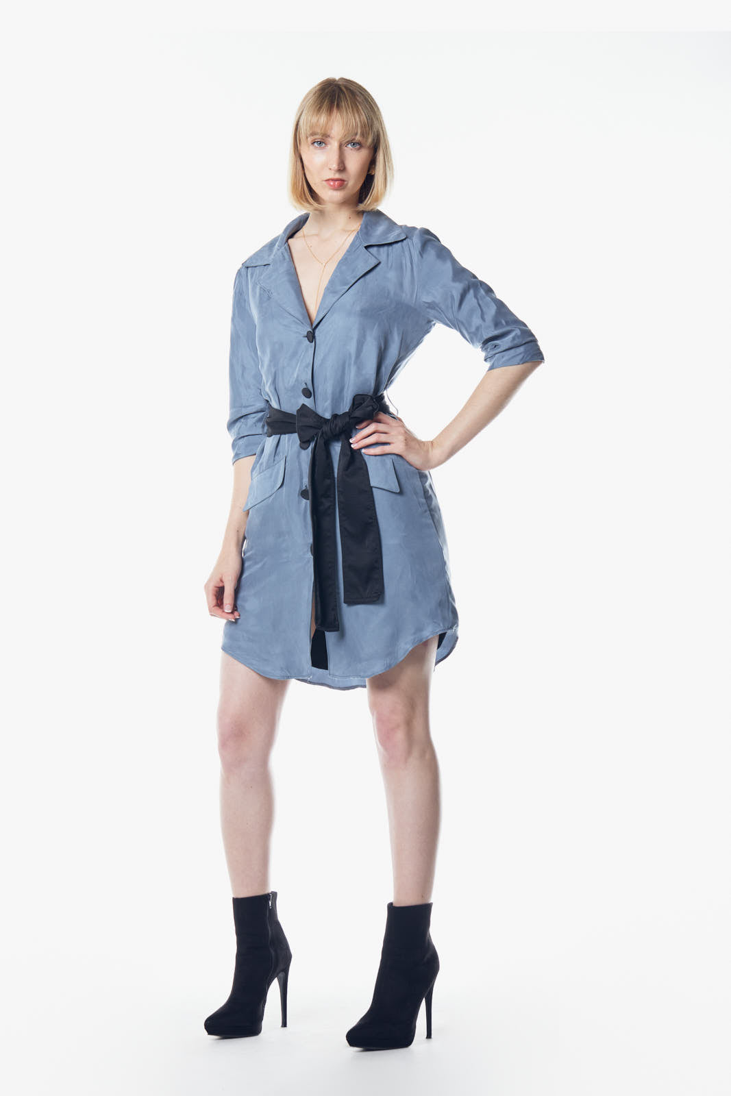 A person stands against a plain background, wearing the Cielo Blue Blazer Dress by Le Réussi®, accented with a black belt and paired with black ankle boots.