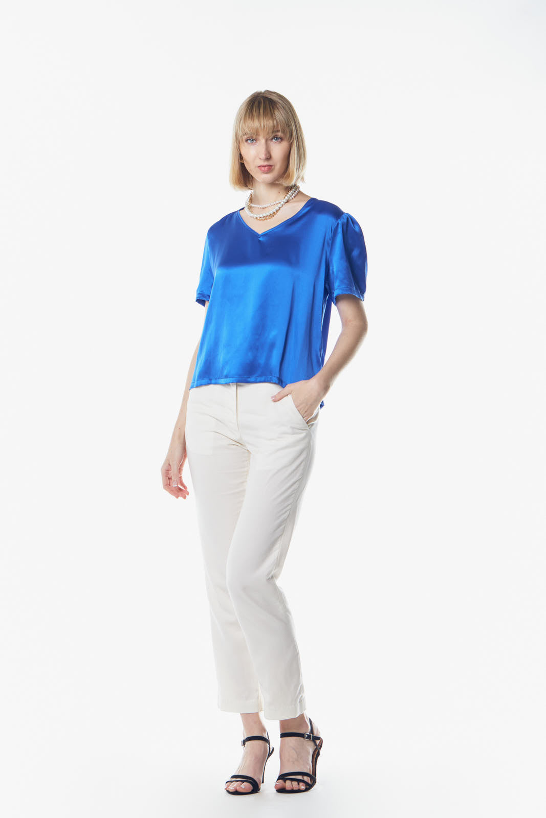 A person with short blonde hair wears a Le Réussi® Shining V-Neck Blouse in Blue and a pearl necklace, standing against a plain white background.