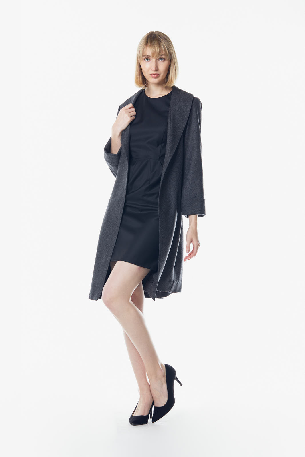 A person poses in a dark dress and the Power Woman Worsted Flannel Long Jacket in Dark Grey by Le Réussi®, against a white background, wearing black high heels.