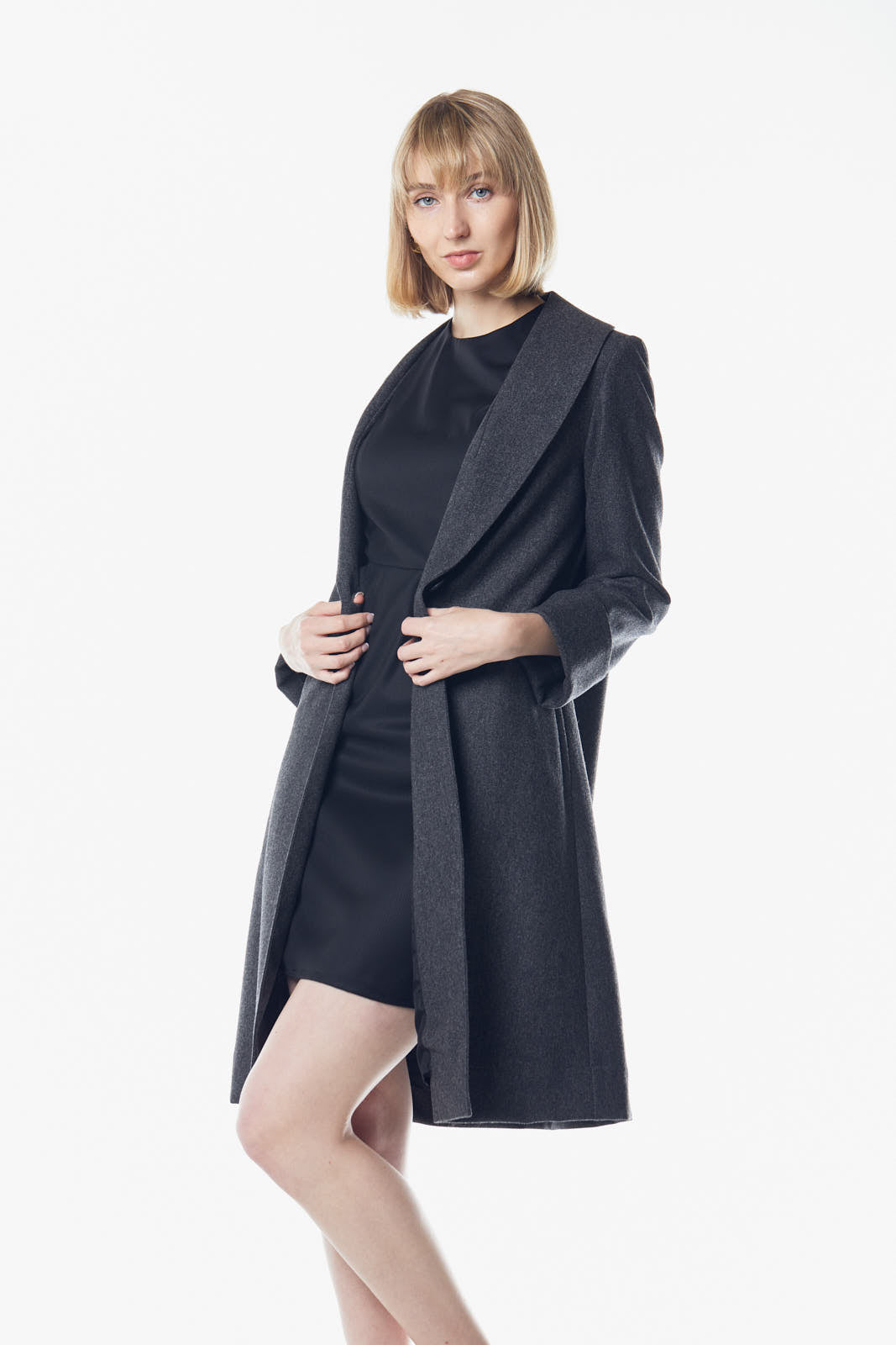 A person poses in a dark dress and the Power Woman Worsted Flannel Long Jacket in Dark Grey by Le Réussi®, against a white background, wearing black high heels.