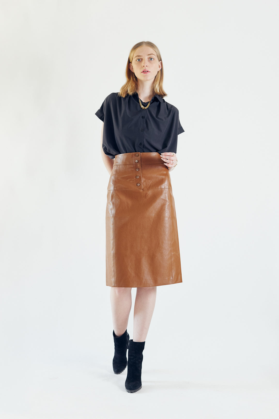 A person wearing a black top, Le Réussi® Power Woman- Dark Hazel Leather Skirt, and black high-heeled boots poses against a plain background.