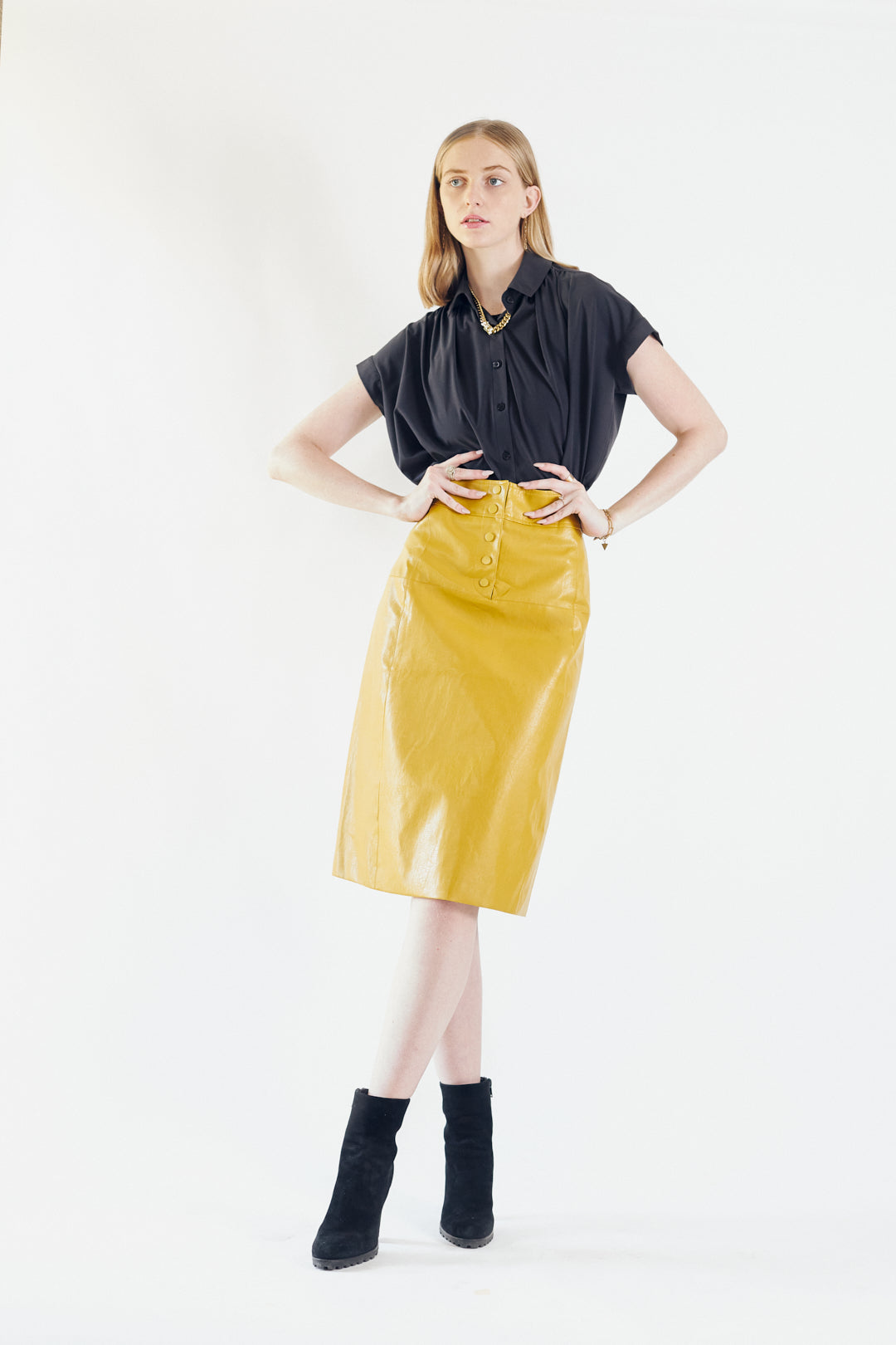 A person is wearing the Power Woman-Mustard Leather Skirt by Le Réussi® and black heeled ankle boots, standing against a white background.