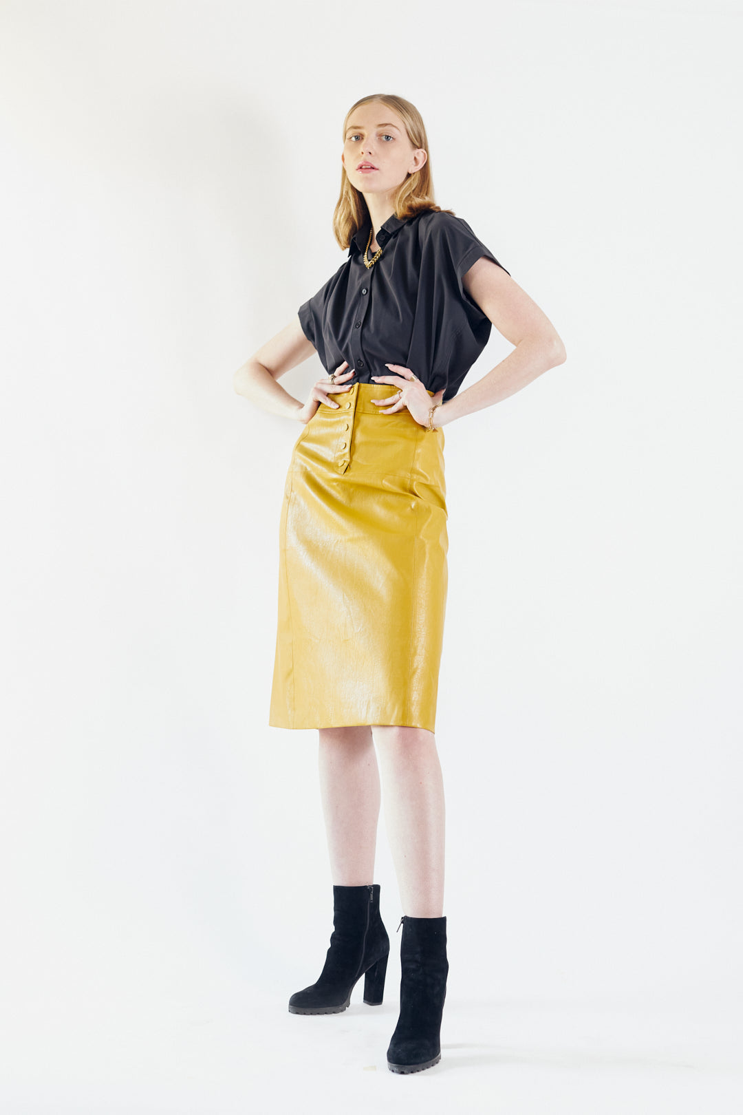 A person is wearing the Power Woman-Mustard Leather Skirt by Le Réussi® and black heeled ankle boots, standing against a white background.