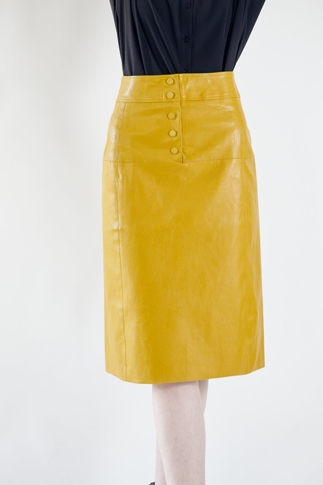 A person is wearing the Power Woman-Mustard Leather Skirt by Le Réussi® and black heeled ankle boots, standing against a white background.