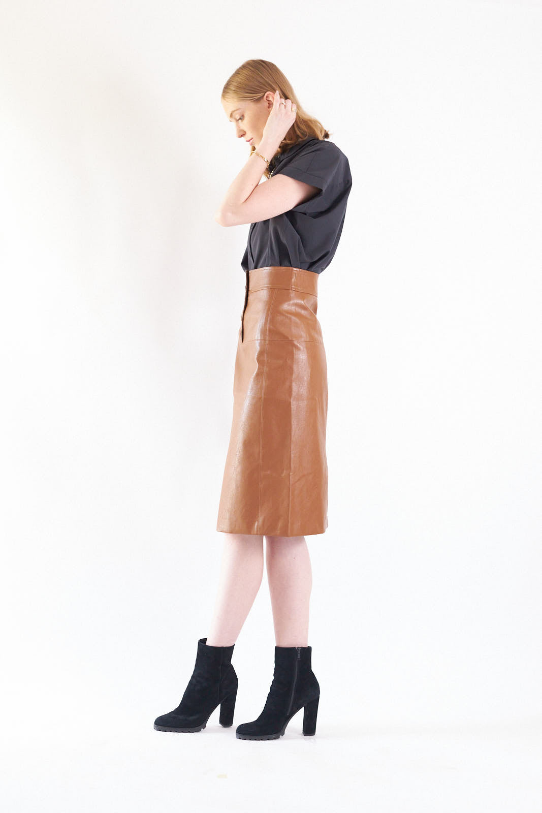 A person wearing a black top, Le Réussi® Power Woman- Dark Hazel Leather Skirt, and black high-heeled boots poses against a plain background.