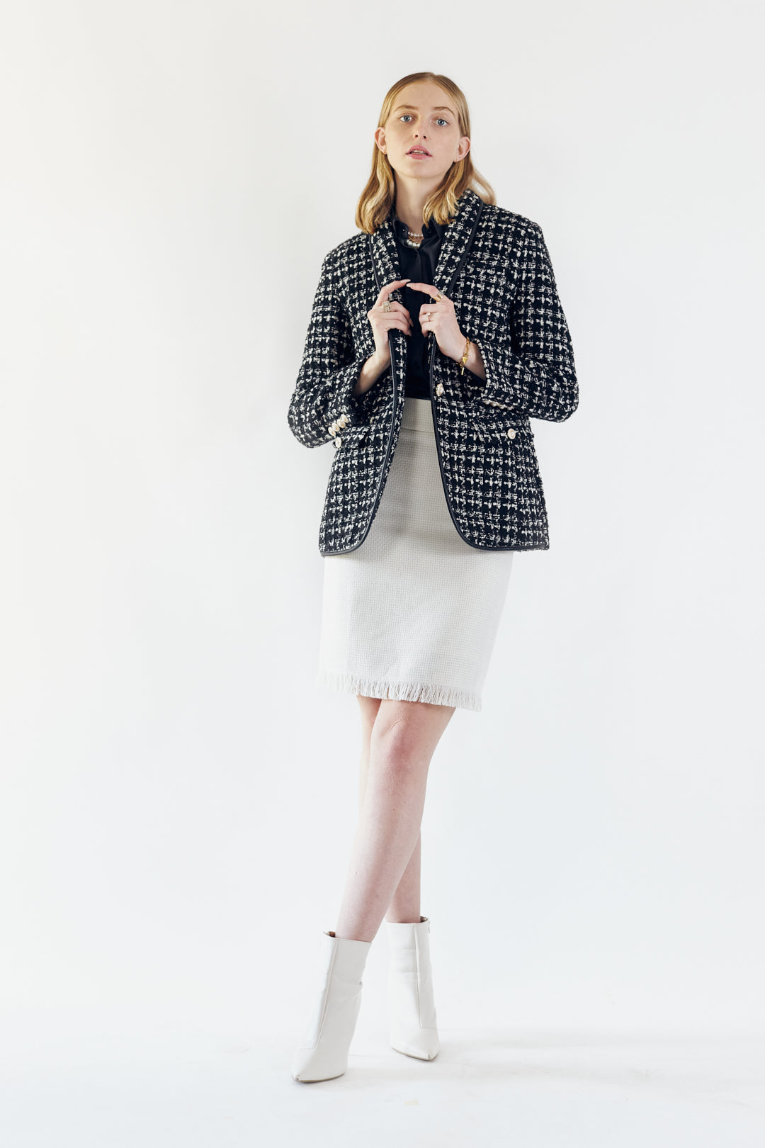A person stands against a plain background wearing a black shirt, Le Réussi® Power Woman- White Tweed Skirt, and white ankle boots.
