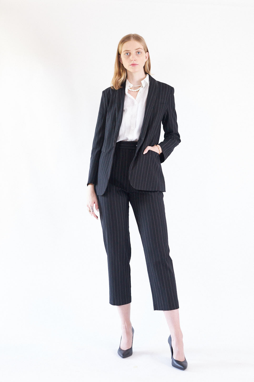 A woman exudes confidence in Le Réussi®s Power Woman- Black/White Stripes Pants paired with a white blouse, standing stylishly with one hand in her pocket against a white background.