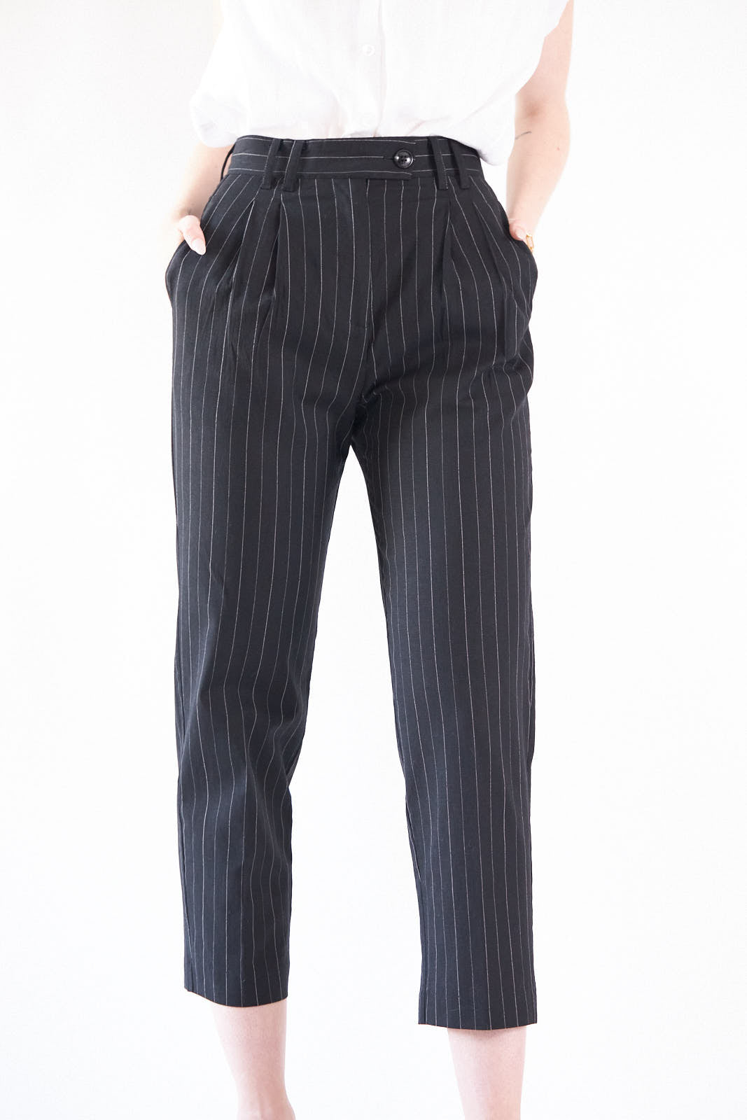A woman exudes confidence in Le Réussi®s Power Woman- Black/White Stripes Pants paired with a white blouse, standing stylishly with one hand in her pocket against a white background.